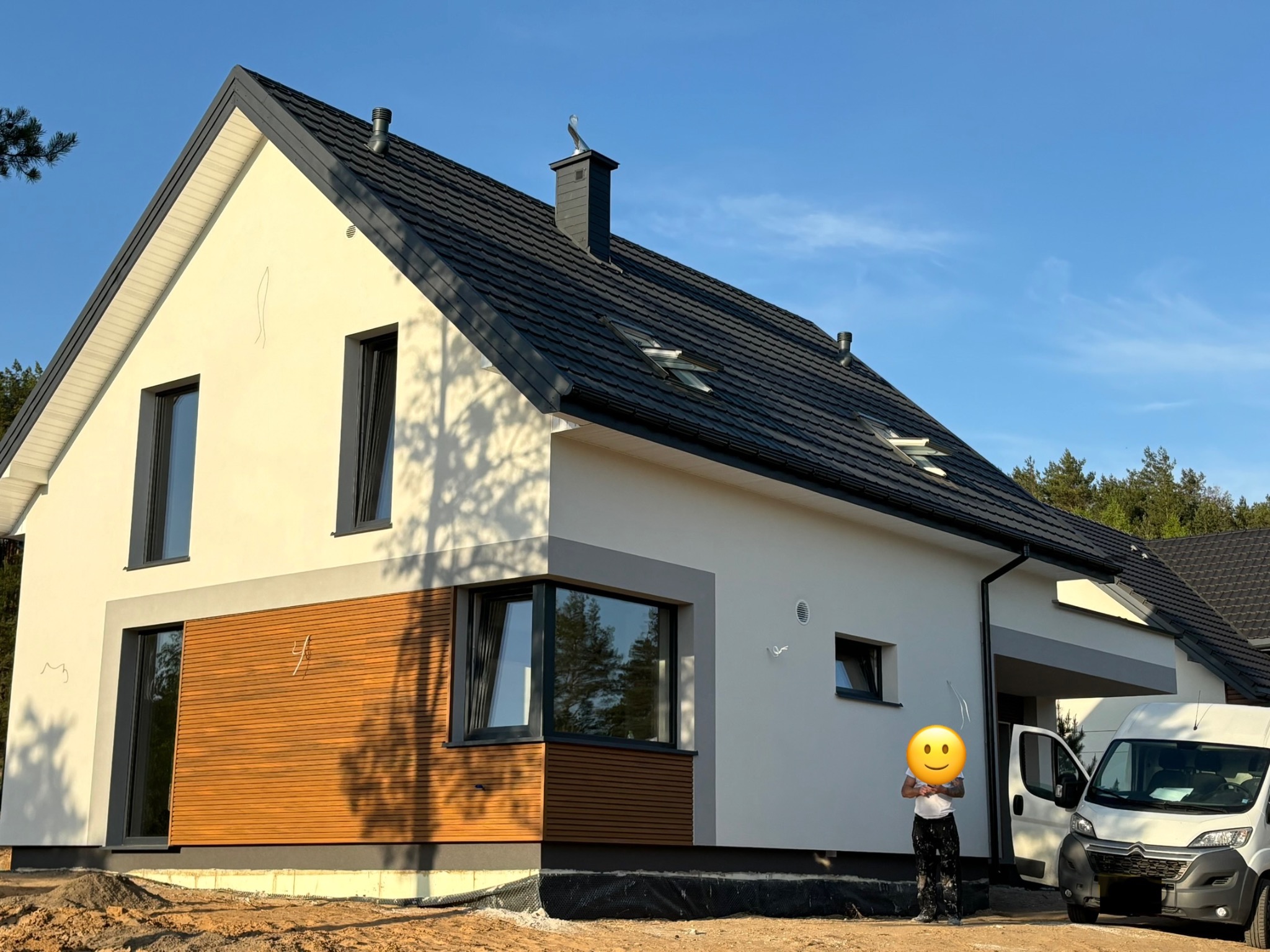 Nowoczesny dom jednorodzinny z drewnianą elewacją i ciemną dachówką, widziany z perspektywy ziemi, z samochodem dostawczym i osobą w tle.