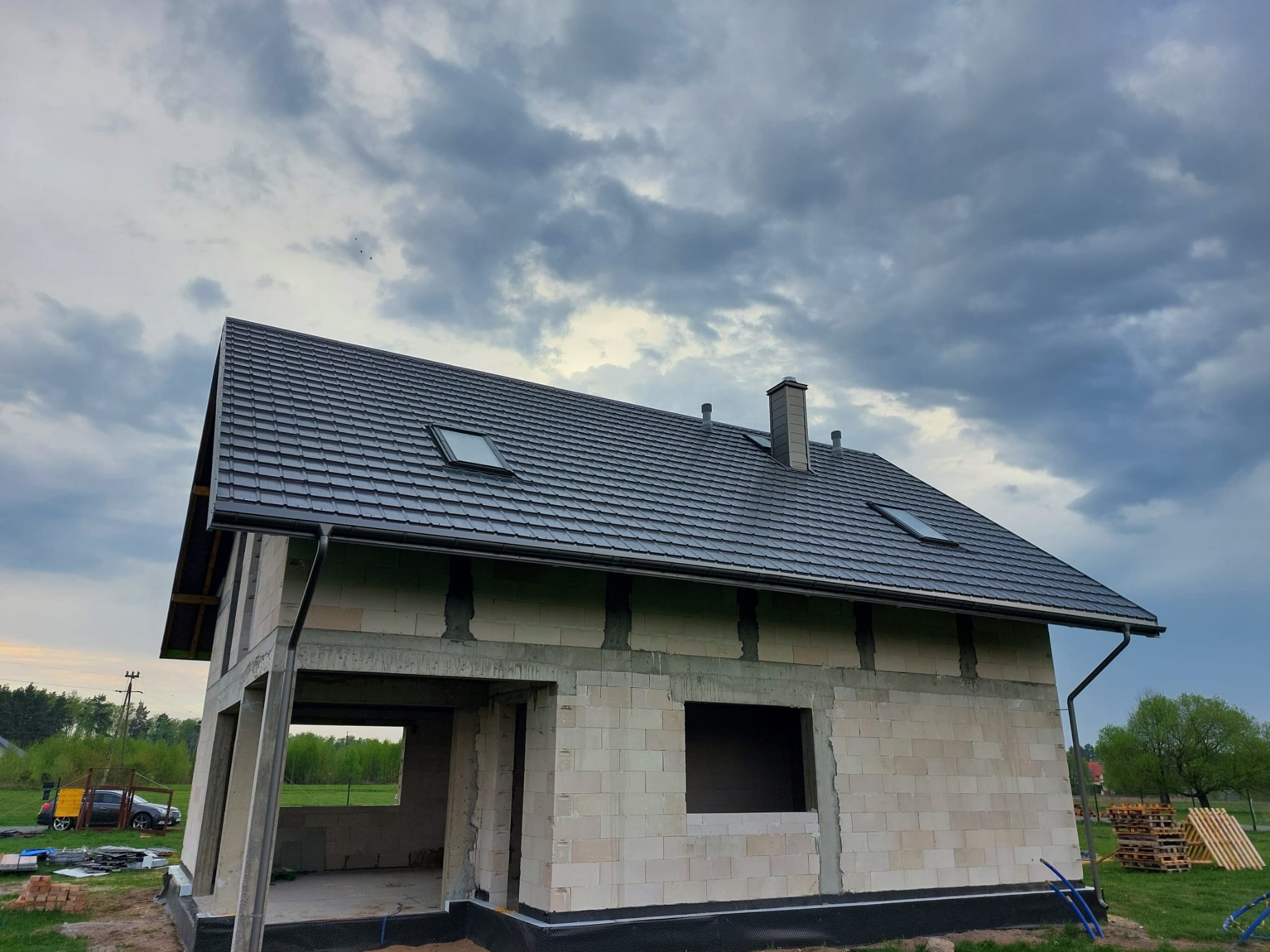 Surowy dom jednorodzinny z szarej cegły silikatowej pod ciemnym dachem z oknami połaciowymi, na tle pochmurnego nieba. Widoczne palety i elementy budowlane.