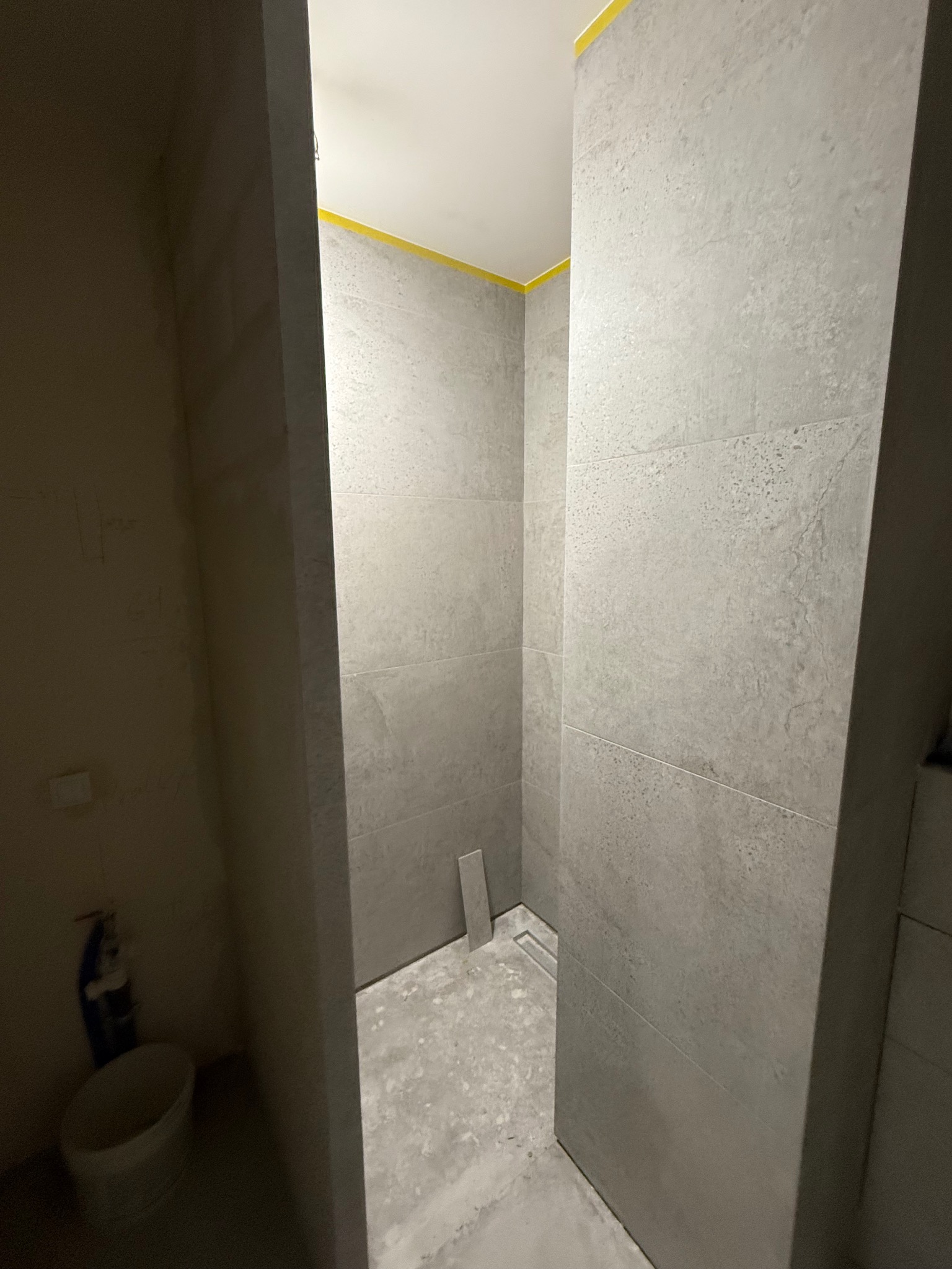 Wykończona w szarościach kabina prysznicowa z odpływem liniowym, zabezpieczona żółtą taśmą malarską przy suficie, w trakcie prac wykończeniowych w łazience.