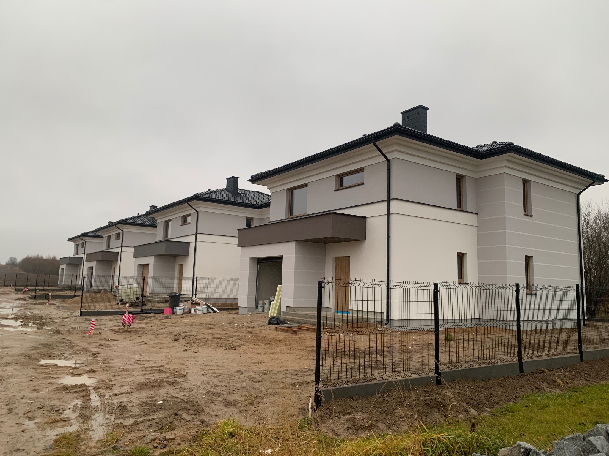 Rząd nowo wybudowanych, dwukondygnacyjnych domów z szarymi elewacjami i czarnymi dachami, otoczonych błotnistym terenem i ogrodzeniem z siatki.