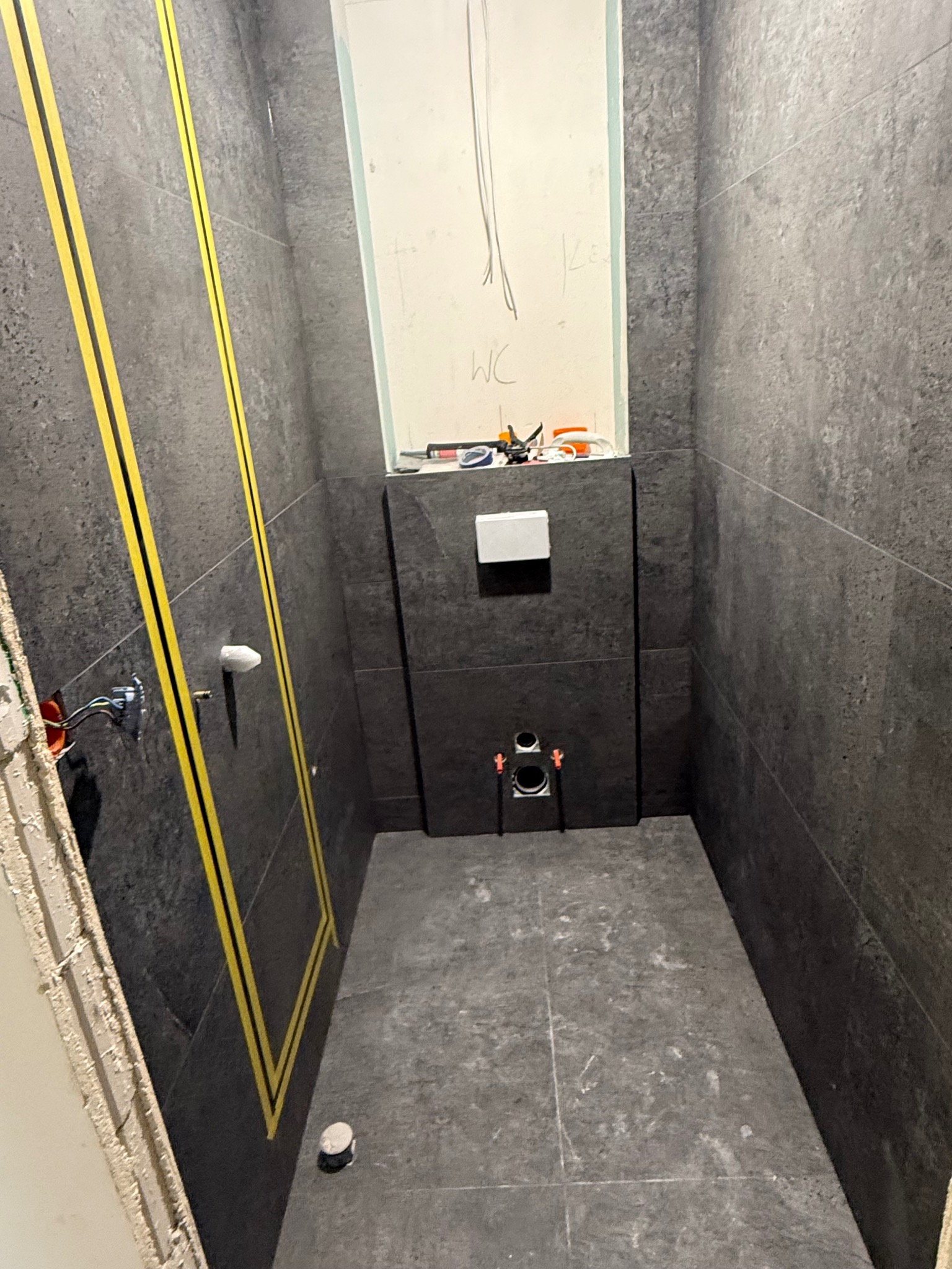 Ciemna łazienka w trakcie remontu: szare płytki, żółte taśmy wyznaczające obszar, przygotowane instalacje pod WC i umywalkę. Widoczne narzędzia i notatki na ścianie.