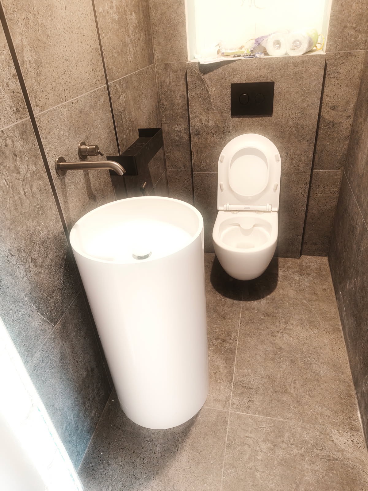 Nowoczesna łazienka z wolnostojącą umywalką i podwieszaną toaletą na tle szarych płytek. Minimalistyczny design, chromowana bateria, czarny panel sterujący.