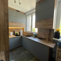 Nowoczesna kuchnia w szarościach i drewnie. Górne szafki sięgają sufitu, a podświetlenie LED dodaje ciepła. Na blacie arbuz i czajnik.