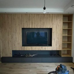MMG MEBLE - Drewniana zabudowa ściany z szafkami, półkami i miejscem na TV. Dekoracyjna wnęka z drewnianymi krążkami. Widoczny odkurzacz i narzędzia.