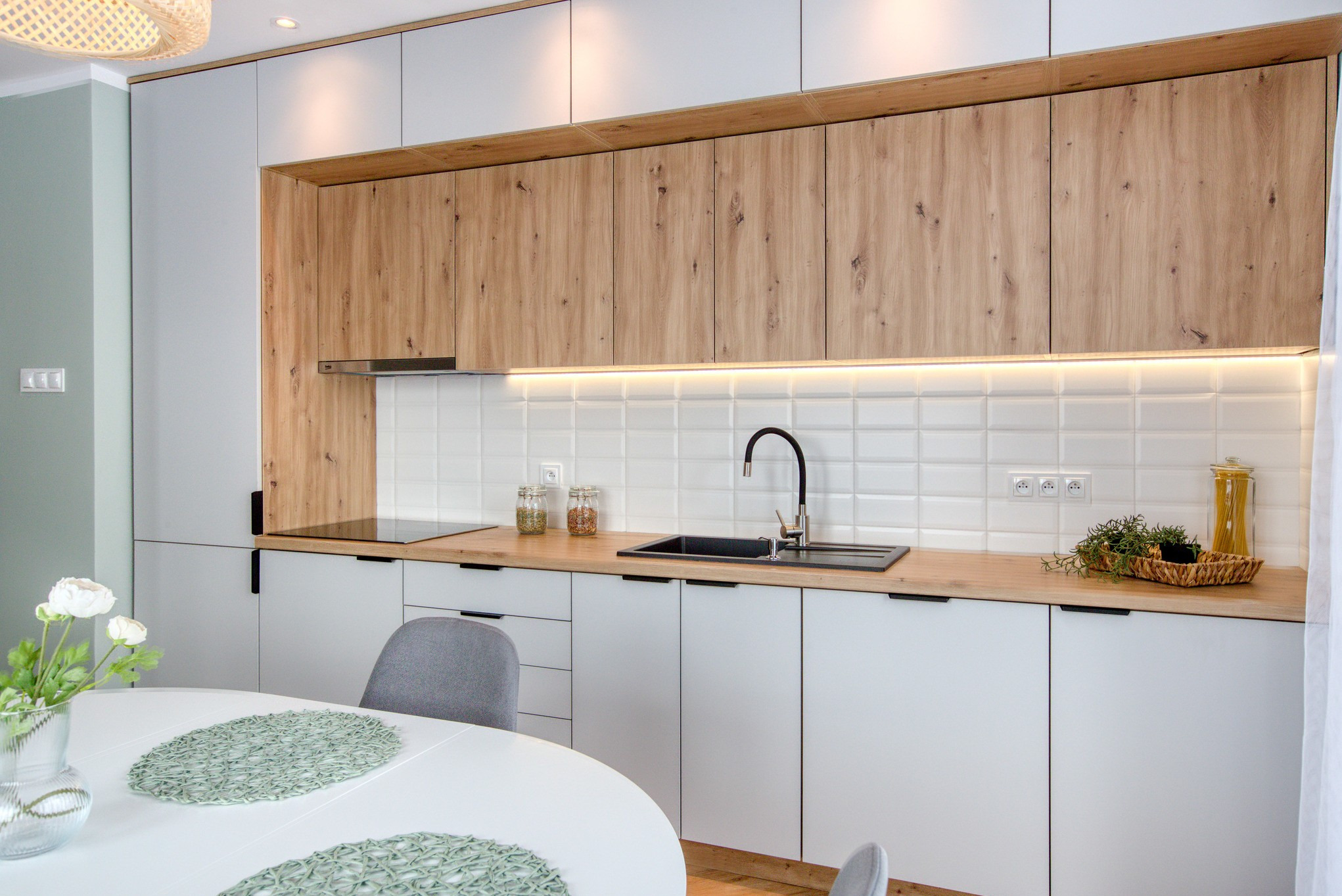 Nowoczesna kuchnia z białymi szafkami dolnymi i górnymi z drewnianymi frontami. Blat drewniany, czarny zlew i bateria. Białe płytki na ścianie i okrągły stół.