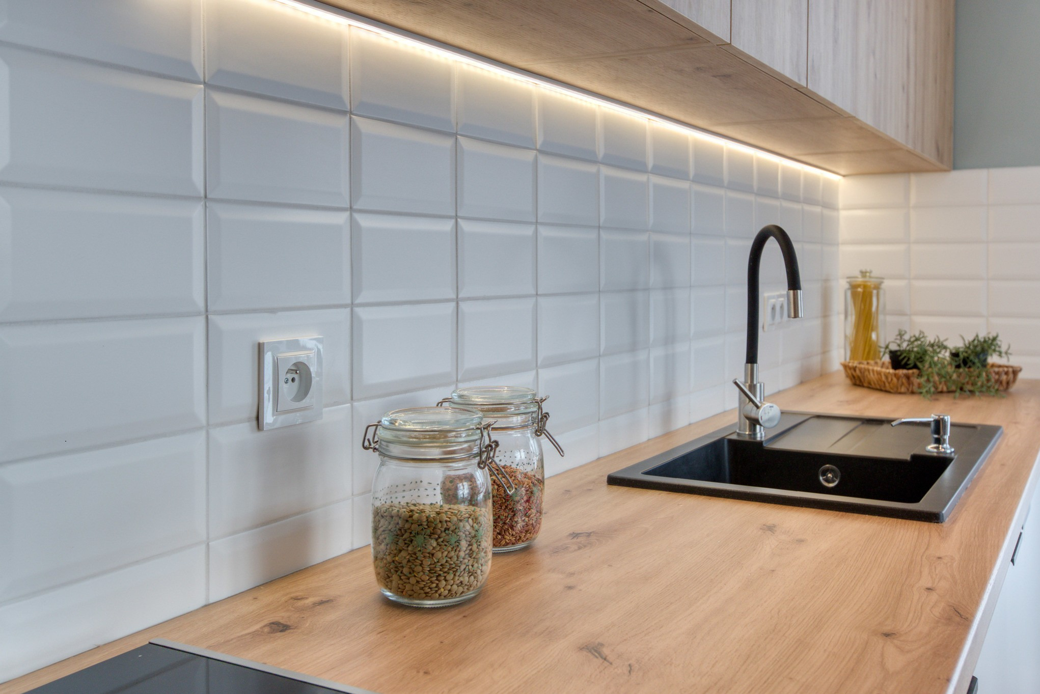 Jasna kuchnia z drewnianym blatem, białymi płytkami i czarnym zlewem. Dwa słoiki z przyprawami na blacie. Oświetlenie LED pod szafkami.