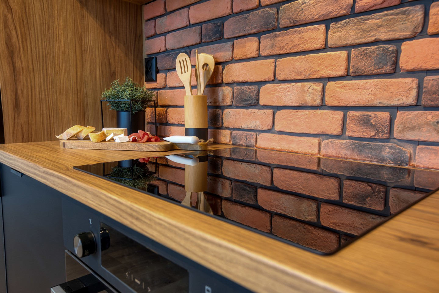 Nowoczesna kuchnia z blatem z drewna, ceglaną ścianą i czarną płytą indukcyjną. Na blacie deska z przekąskami i zioła w doniczce.