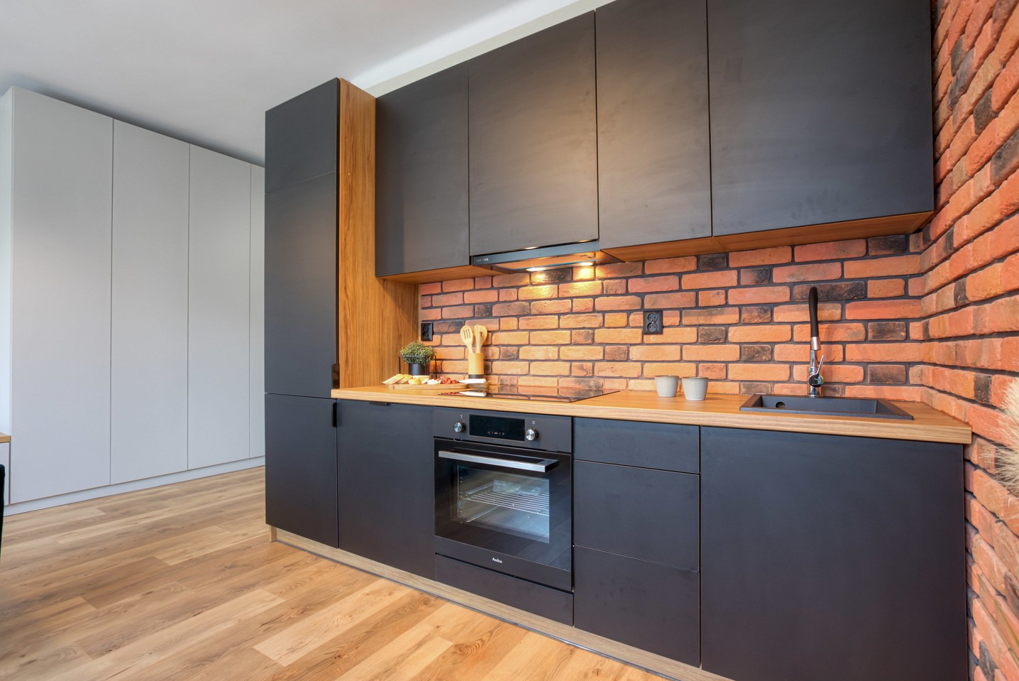 Nowoczesna kuchnia z czarnymi szafkami, drewnianym blatem i ceglaną ścianą. Widoczny piekarnik Amica i drewniane akcenty. Minimalistyczny design.