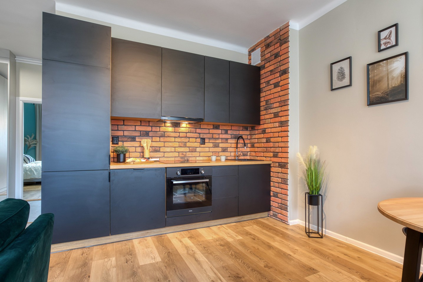 Nowoczesna kuchnia z czarnymi matowymi frontami, drewnianym blatem i ceglaną ścianą. Oświetlenie punktowe podkreśla fakturę cegły. Widoczne detale: roślina, obrazy na ścianie.