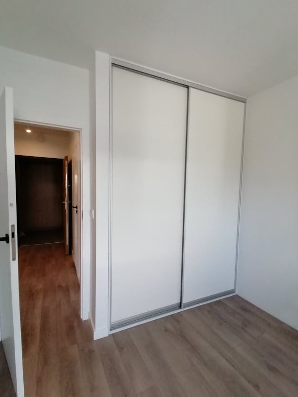 Biała szafa przesuwna z szarymi obramowaniami w minimalistycznym wnętrzu. Widoczna podłoga z paneli i otwarte drzwi do innego pomieszczenia.