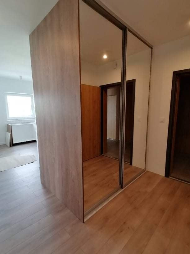 Nowoczesna szafa wnękowa z lustrzanymi drzwiami przesuwnymi, wykończona drewnopodobną okleiną, w jasnym wnętrzu z drewnianą podłogą i widocznymi drzwiami.