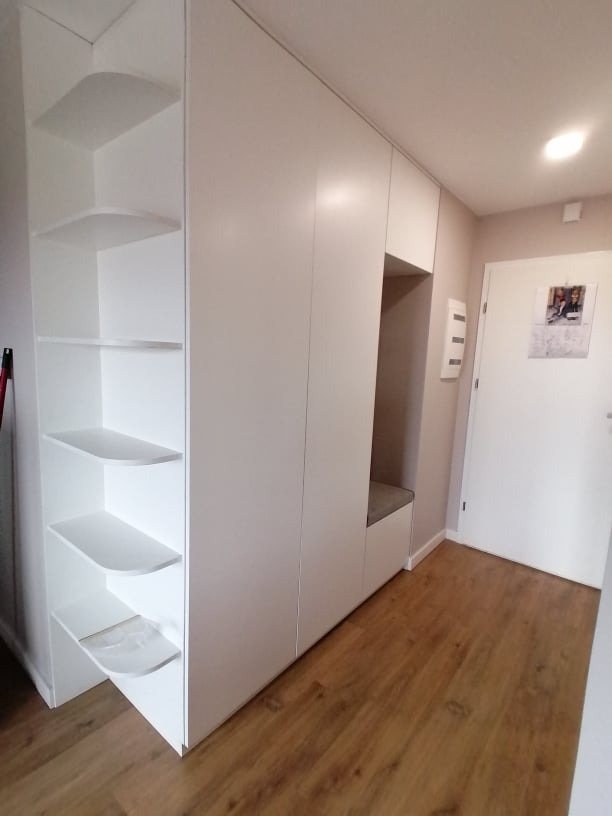 Biała szafa wnękowa z otwartymi półkami i siedziskiem w przedpokoju. Minimalistyczny design, jasne drewno na podłodze, drzwi wejściowe w tle. Funkcjonalne rozwiązanie do przechowywania.