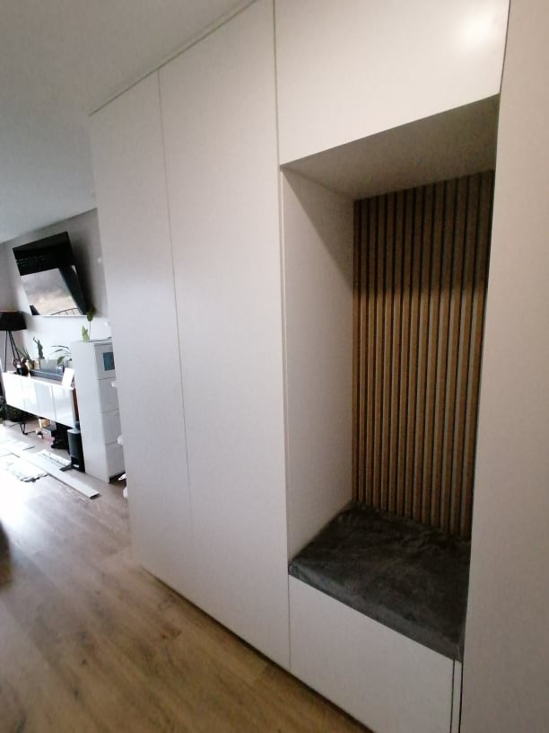 Biała szafa wnękowa z siedziskiem i drewnianymi lamelami na ścianie wnęki, widok z perspektywy podłogi, nowoczesny design.