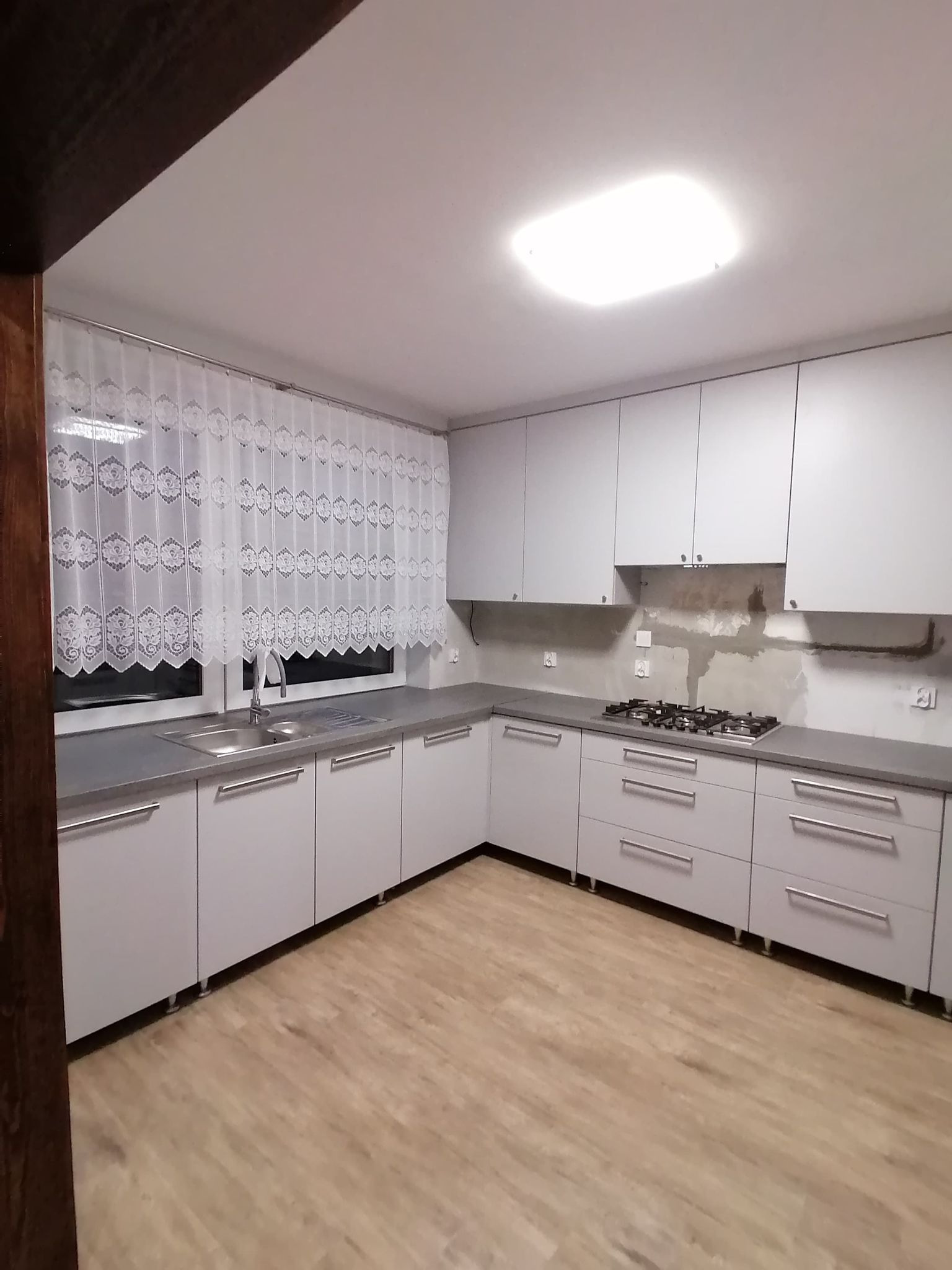 Jasna kuchnia z szarym blatem i drewnianą podłogą. Widoczne białe zasłony w oknie i fragment ściany nad kuchenką w trakcie wykończenia.