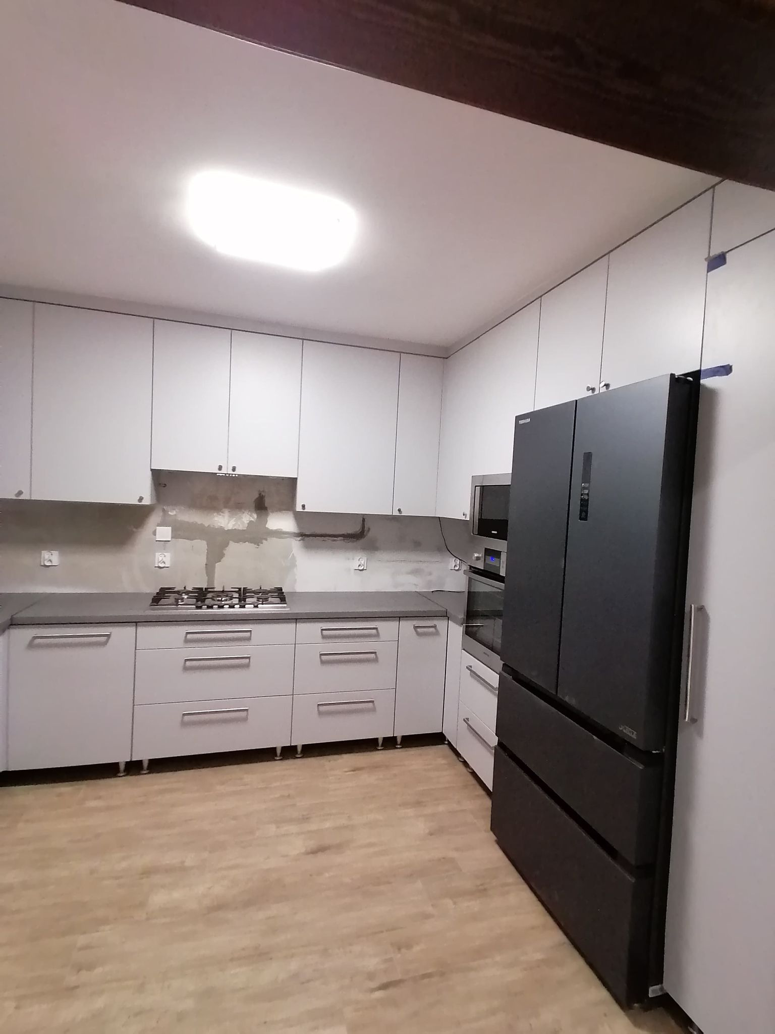 Jasna kuchnia z szarym blatem i czarną lodówką w Będzinie. Widoczne białe szafki wiszące i dolne, płyta gazowa oraz fragment ściany przygotowanej pod montaż paneli.