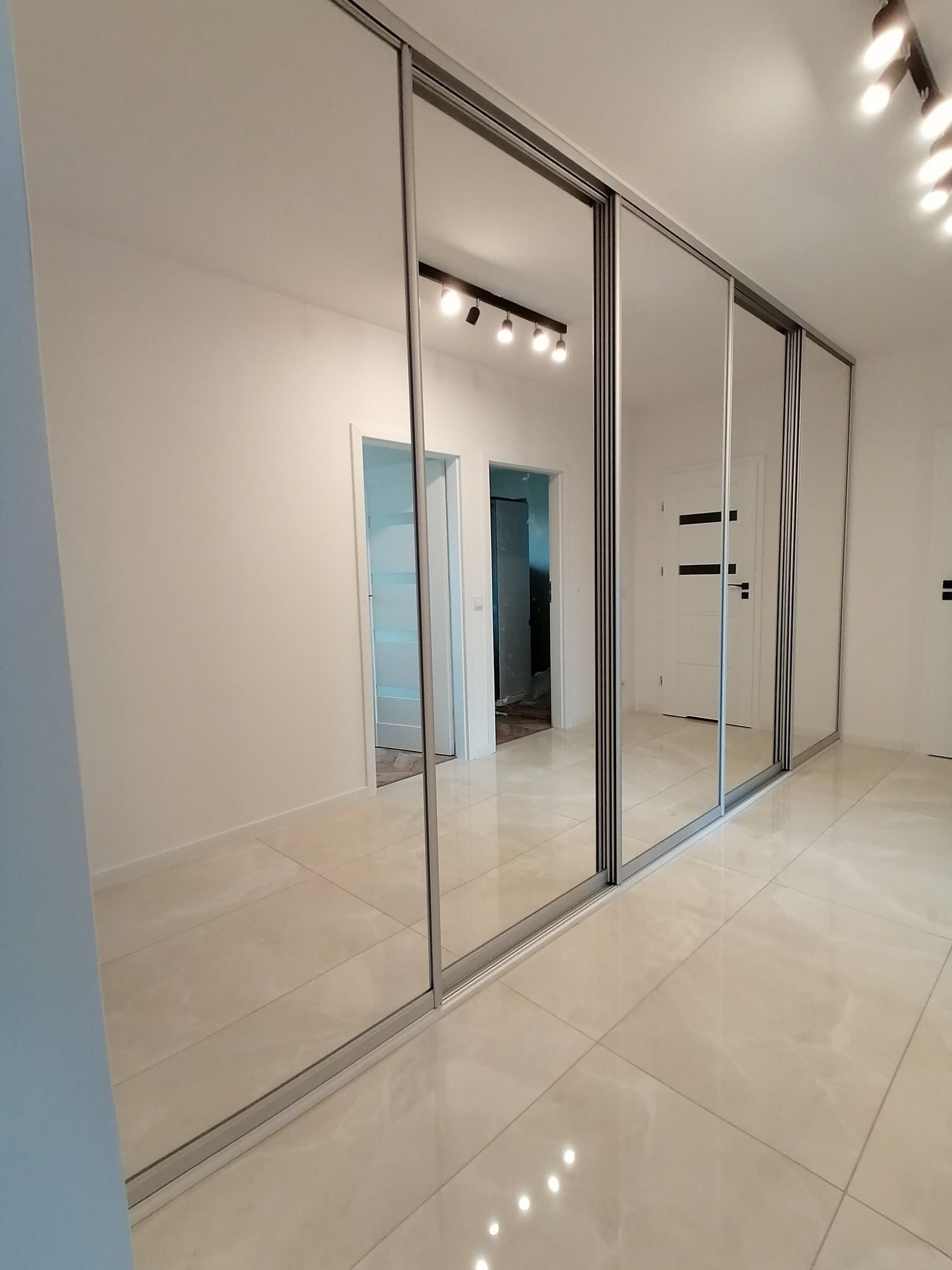 Nowoczesna szafa wnękowa z lustrzanymi drzwiami przesuwnymi, odbijająca wnętrze jasnego pomieszczenia z białymi ścianami i podłogą z płytek.