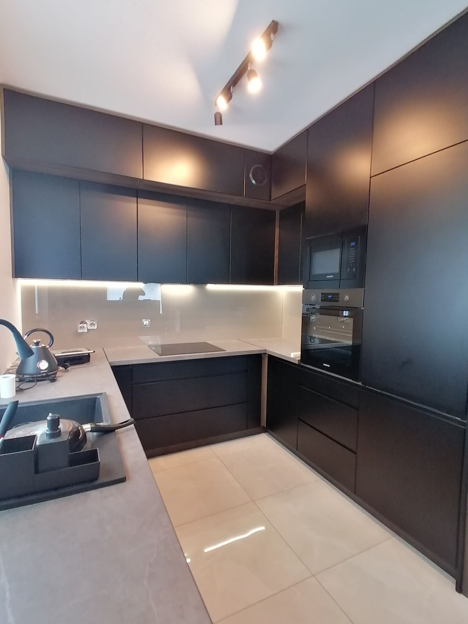 Nowoczesna, czarna kuchnia z matowymi frontami, oświetleniem LED pod szafkami i beżowymi płytkami podłogowymi. Widoczny czajnik i płyta indukcyjna.