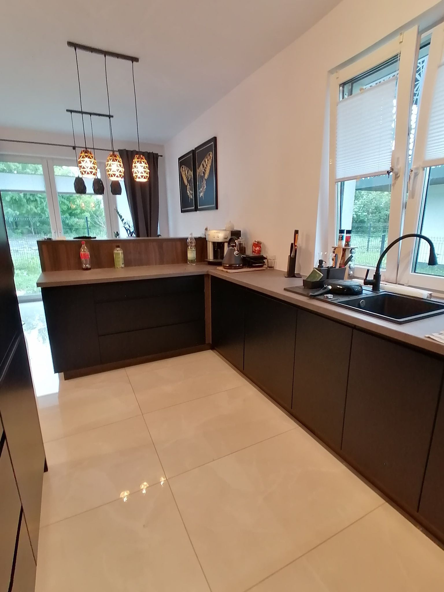 Nowoczesna kuchnia z czarnymi frontami, drewnianym blatem i designerskimi lampami wiszącymi. Widok na okno z żaluzjami i dekoracje ścienne.