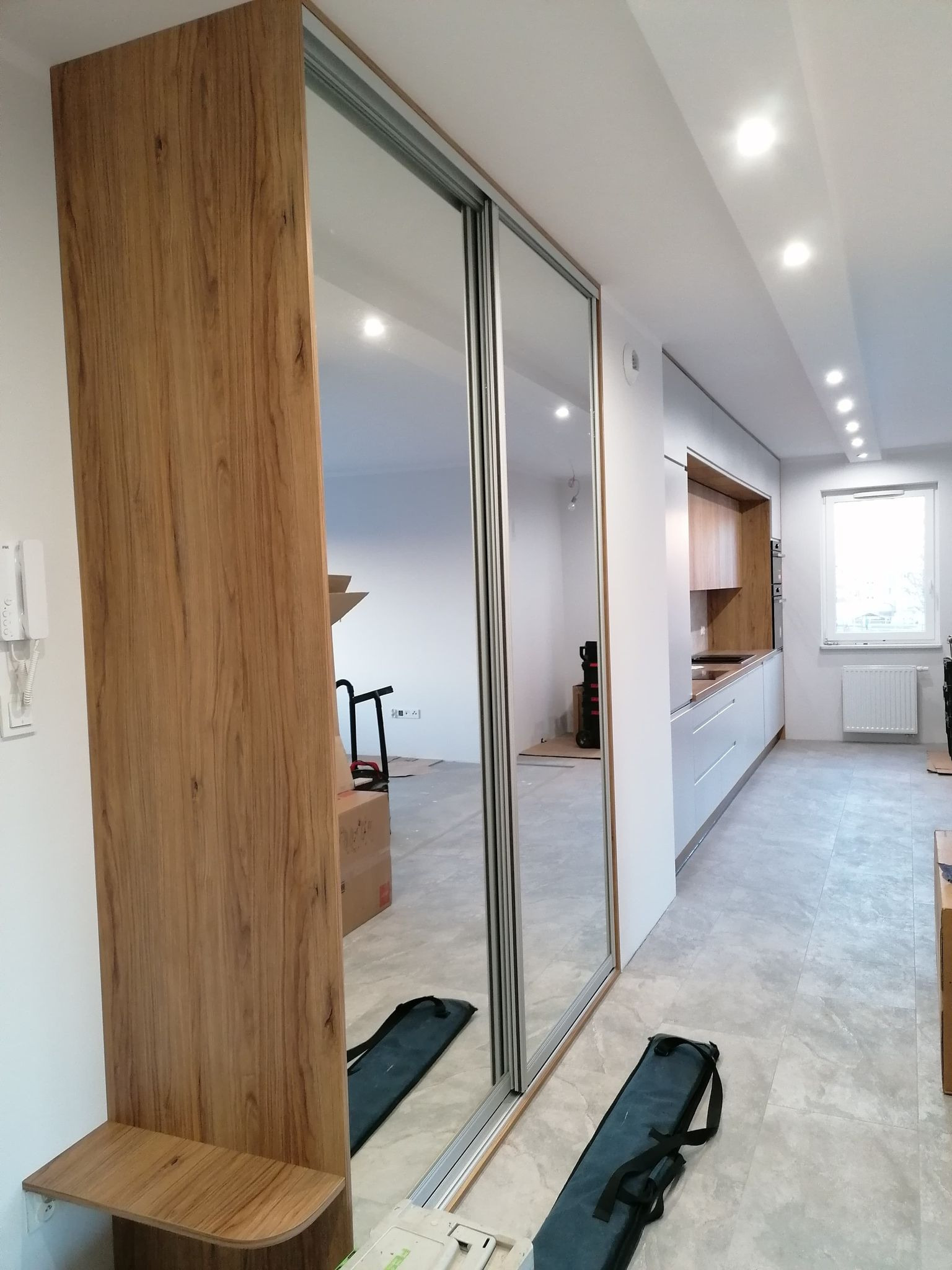 Nowoczesna szafa wnękowa z lustrzanymi drzwiami przesuwnymi i drewnianym panelem bocznym, widoczna w jasnym wnętrzu mieszkania z elementami zabudowy kuchennej.