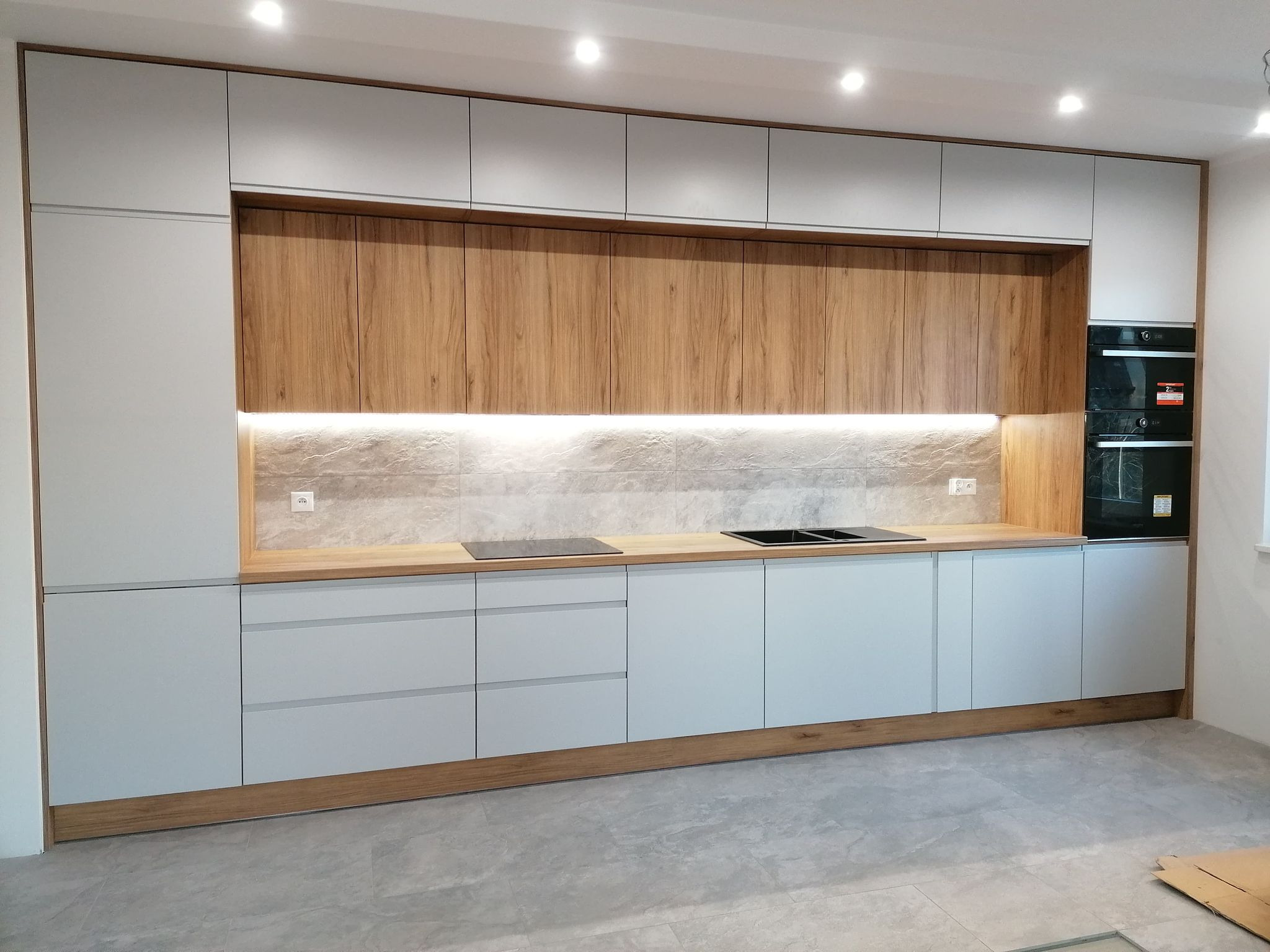 Nowoczesna kuchnia z szarymi frontami i drewnianymi akcentami. Górne szafki podświetlone, płyta indukcyjna i piekarnik w zabudowie. Minimalistyczny design.