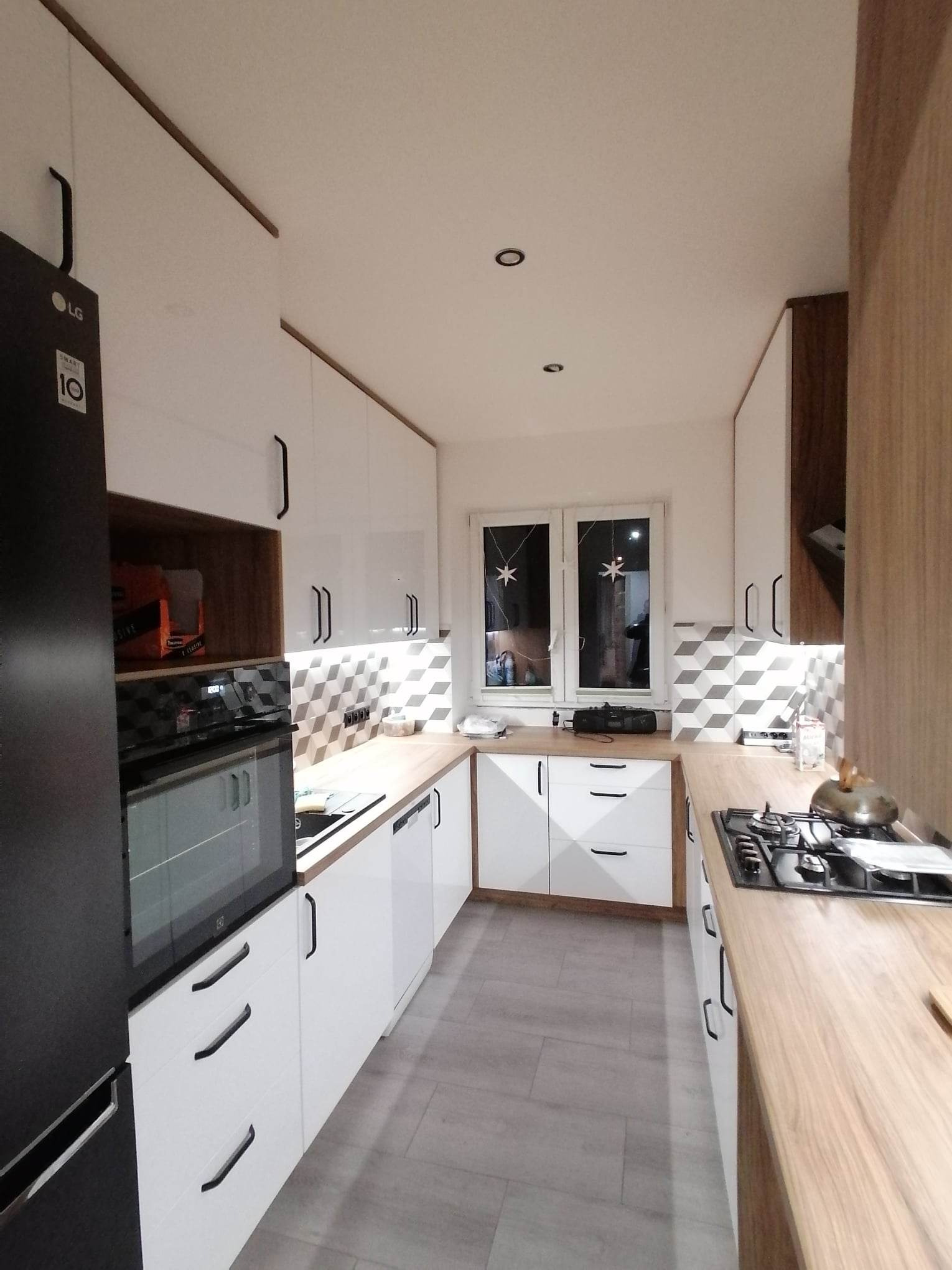 Nowoczesna kuchnia z białymi, lakierowanymi frontami, drewnianymi blatami i geometrycznym wzorem na ścianie. Widoczny czarny sprzęt AGD i okno z dekoracjami.