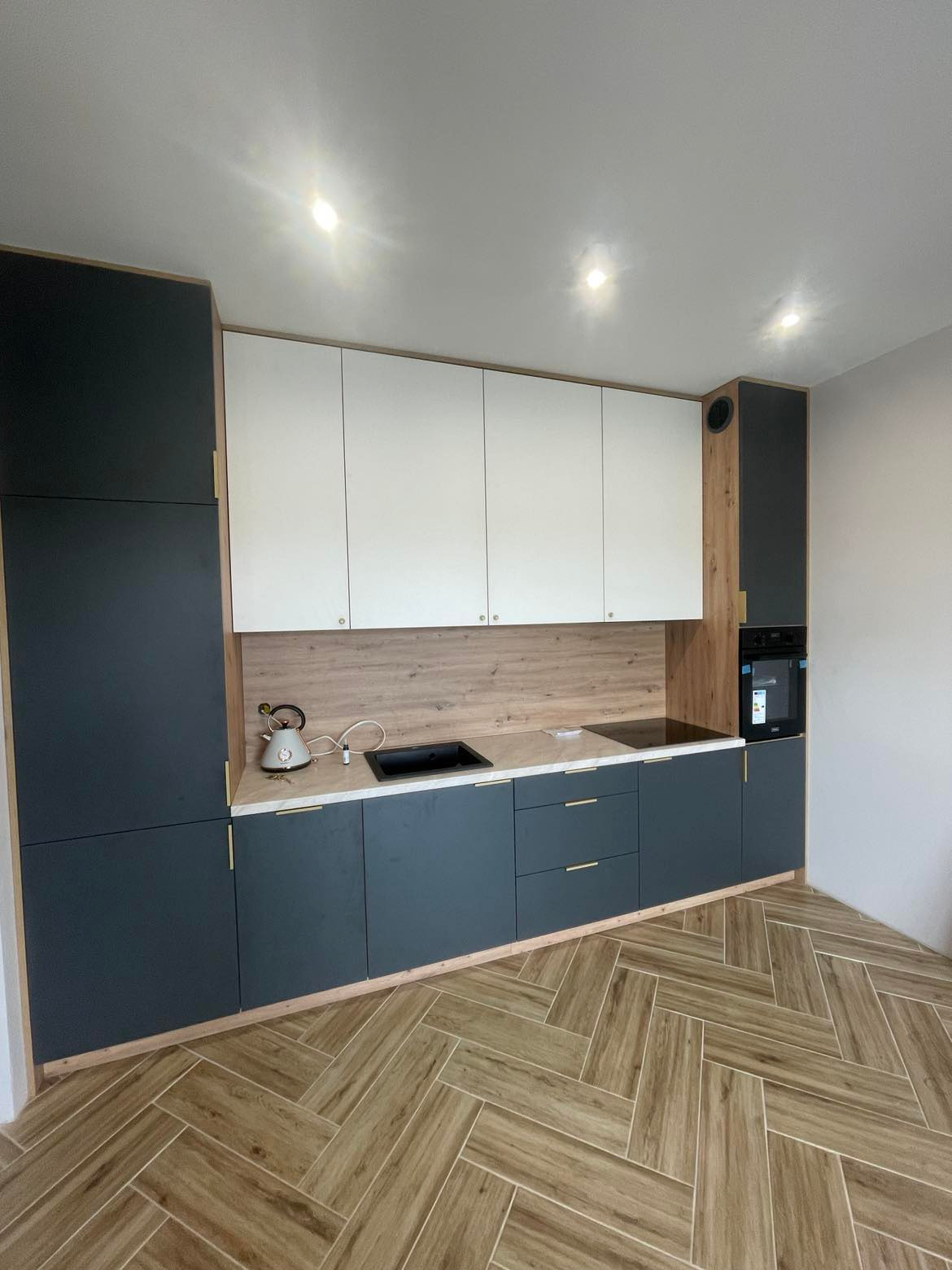 Nowoczesna kuchnia z ciemnymi szafkami dolnymi i białymi górnymi, drewniany panel nad blatem, parkiet w jodełkę. Minimalistyczny design z czajnikiem na blacie.