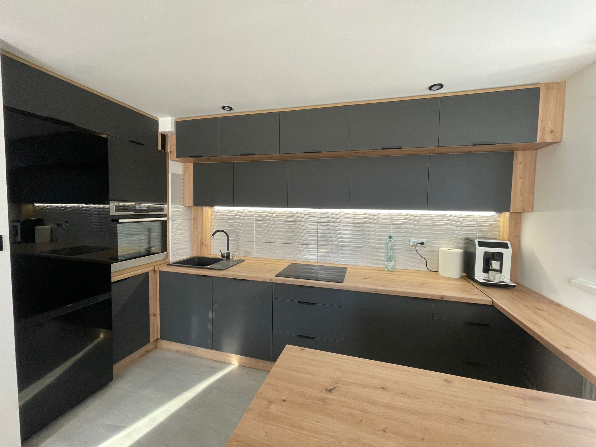 Nowoczesna kuchnia w odcieniach szarości i drewna, z oświetleniem LED pod szafkami wiszącymi i sprzętem AGD. Minimalistyczny design z blatem imitującym drewno.