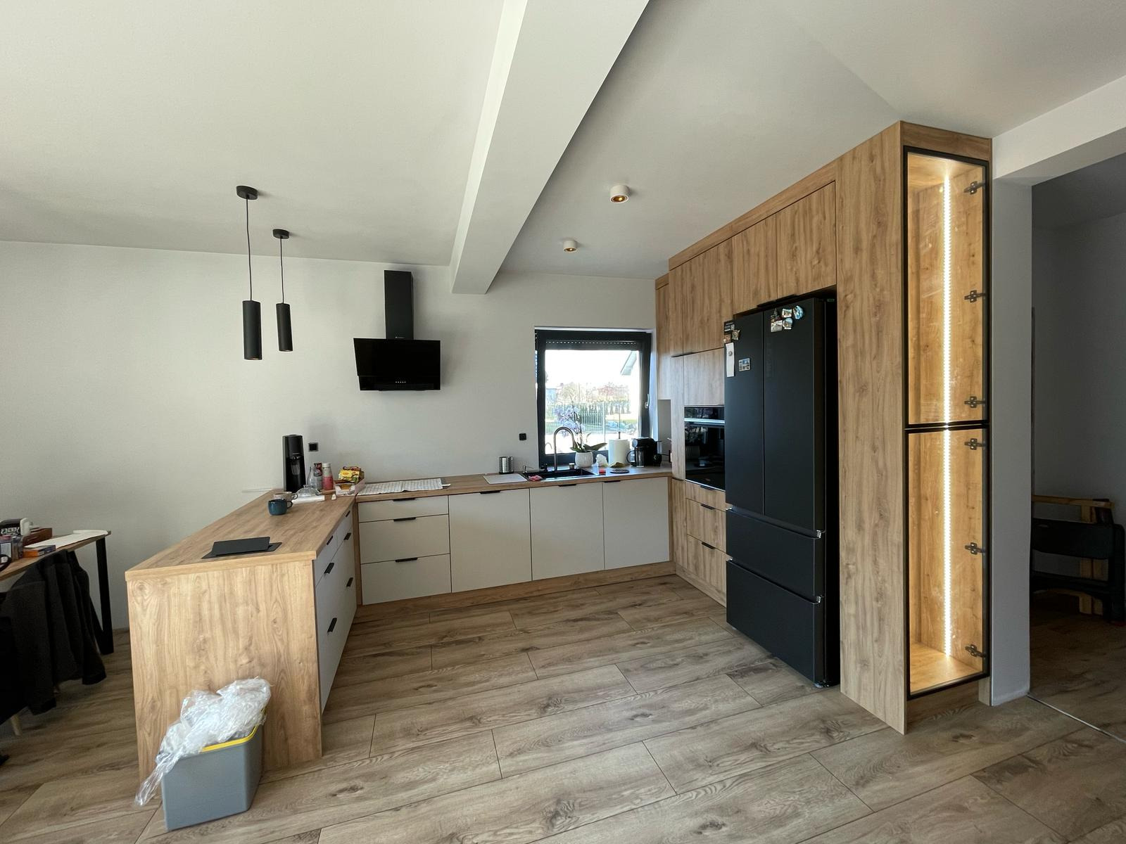 Nowoczesna kuchnia z drewnianymi i białymi frontami, czarnym sprzętem AGD i oświetloną szafką. Widok na drewnianą podłogę i wyspę kuchenną.