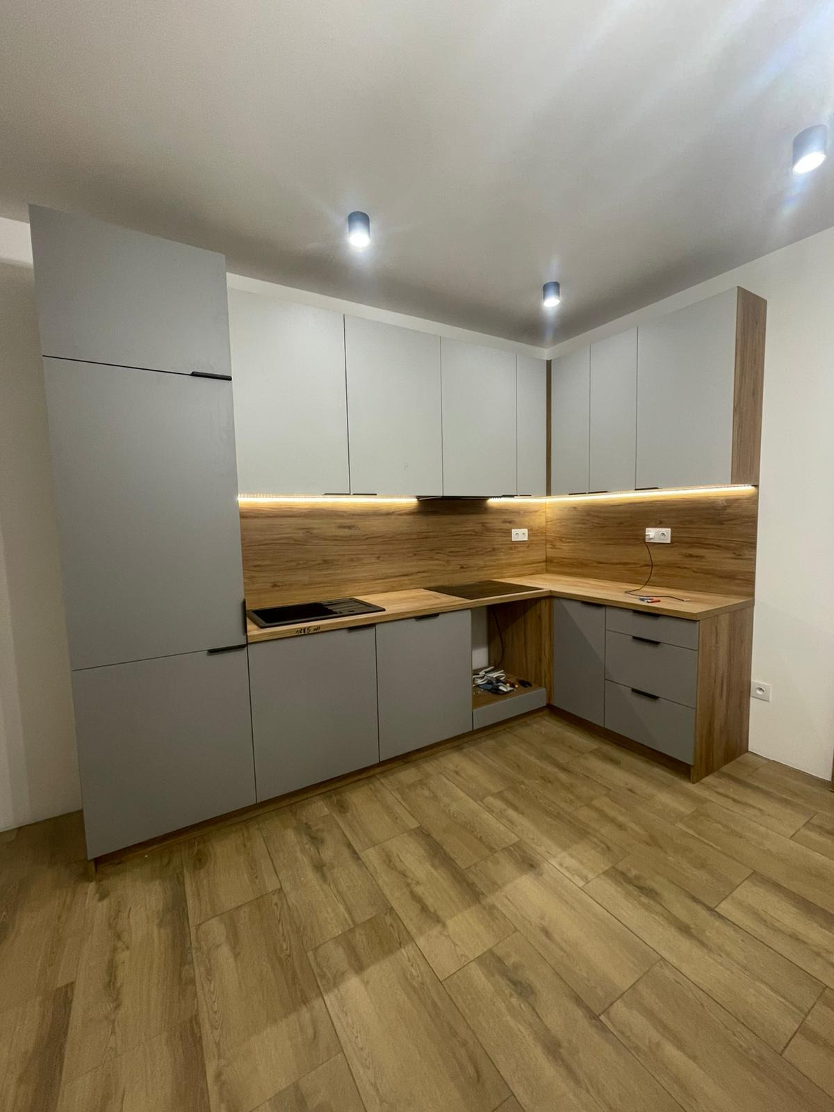 Szara kuchnia na wymiar z drewnianym blatem i oświetleniem LED w Będzinie. Nowoczesny design, minimalistyczne uchwyty, płyta indukcyjna. Widok na całą zabudowę kuchenną.