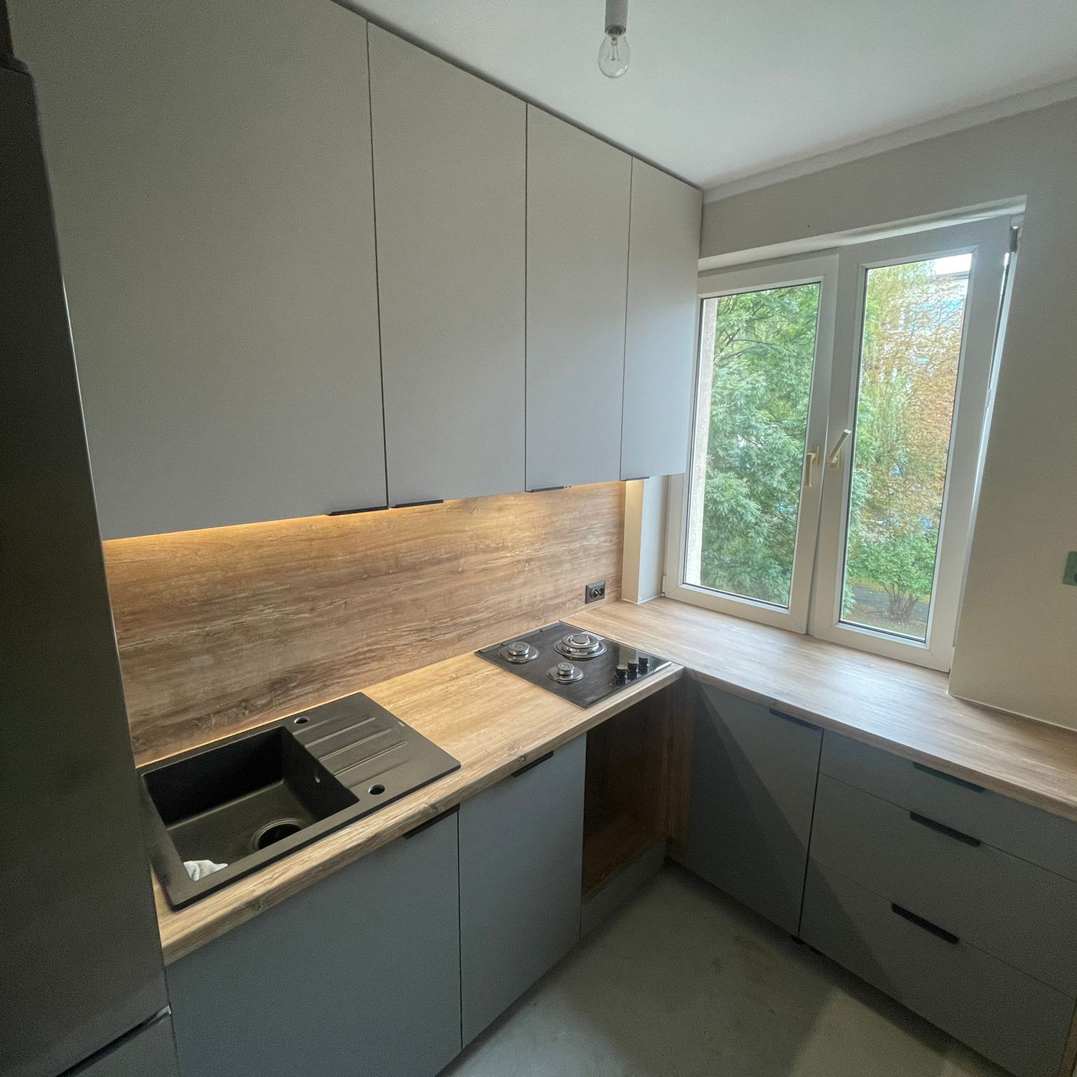 Szara kuchnia z drewnianym blatem i oświetleniem LED pod szafkami wiszącymi. Widok na okno z zielenią. Minimalistyczny design, nowoczesne wykończenie.