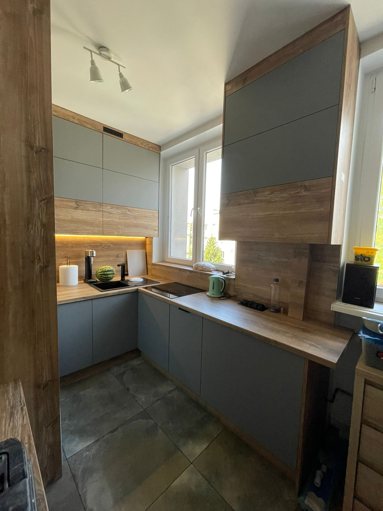 Nowoczesna kuchnia w szarościach i drewnie. Górne szafki sięgają sufitu, a podświetlenie LED dodaje ciepła. Na blacie arbuz i czajnik.