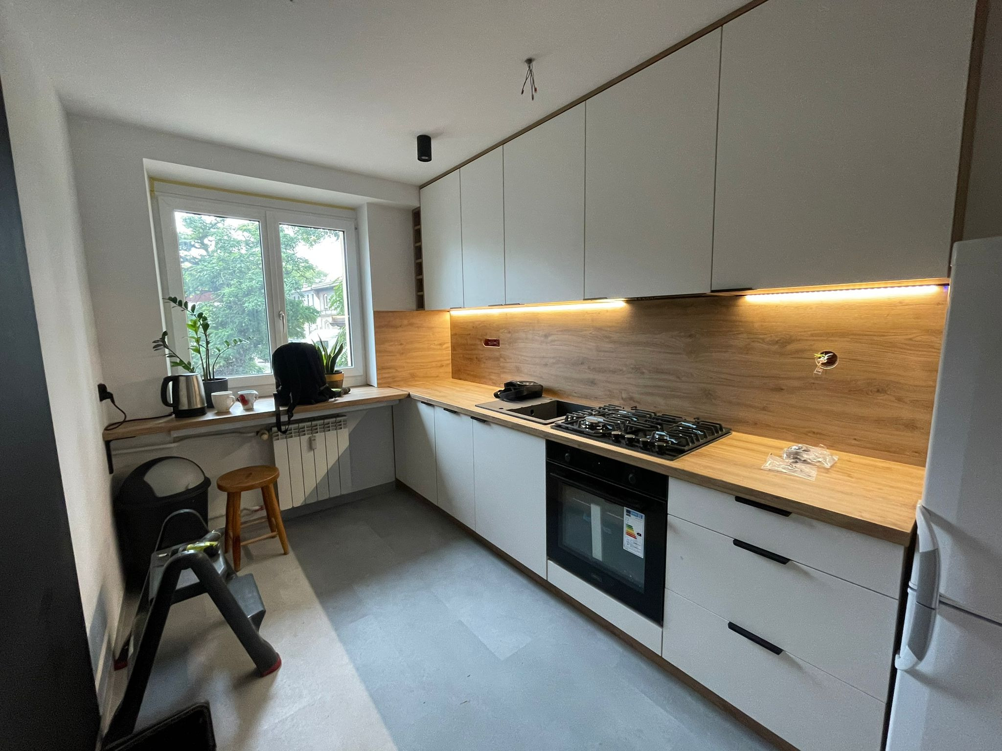 Jasna kuchnia z drewnianym blatem i oświetleniem LED, widok na okno z roślinami. W tle drabina i stołek, sugerujące świeży montaż.