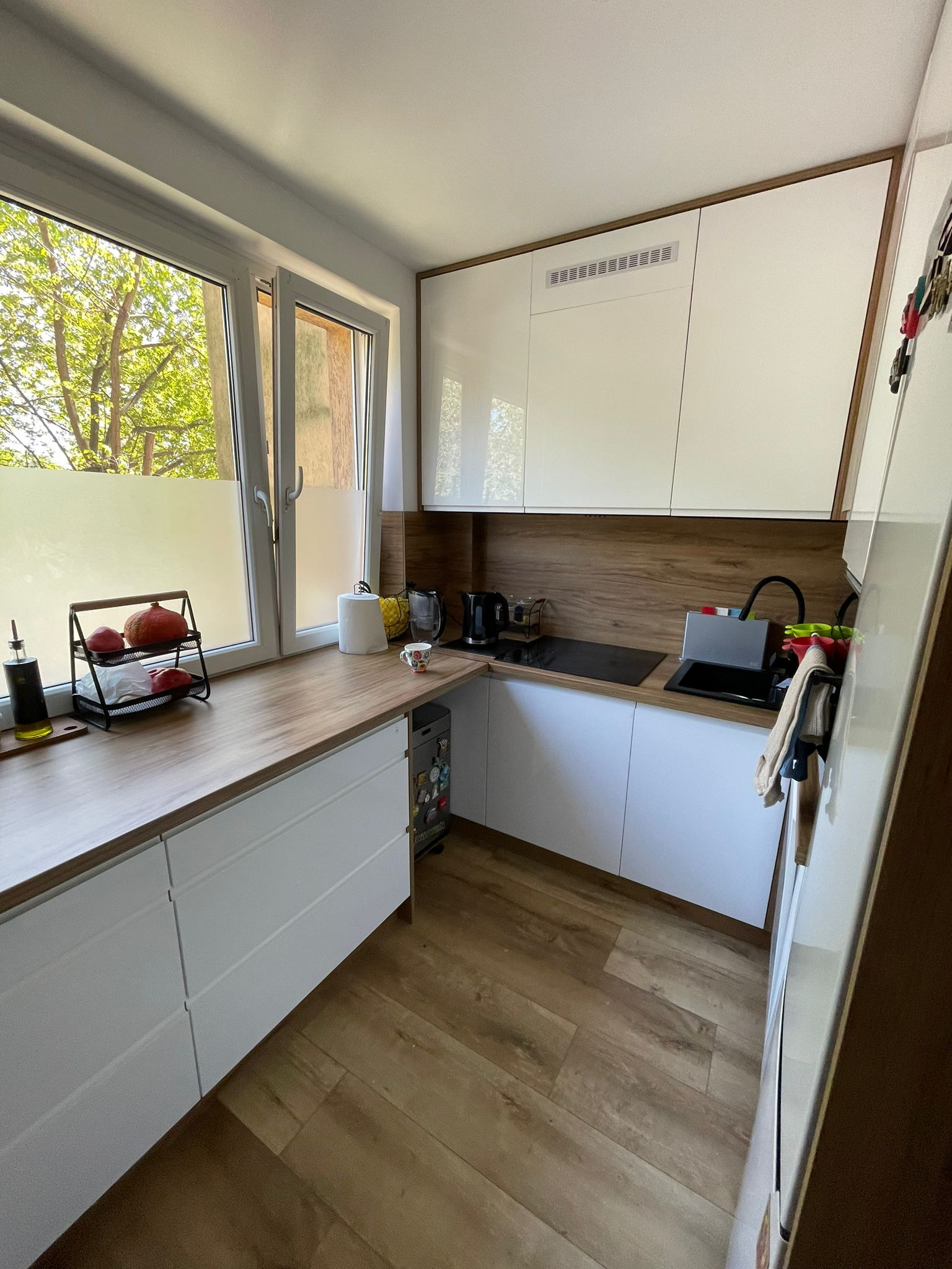 Jasna kuchnia z drewnianym blatem i białymi szafkami, widok na okno z zielenią. Nowoczesny design, czarne akcenty, drewniana podłoga.