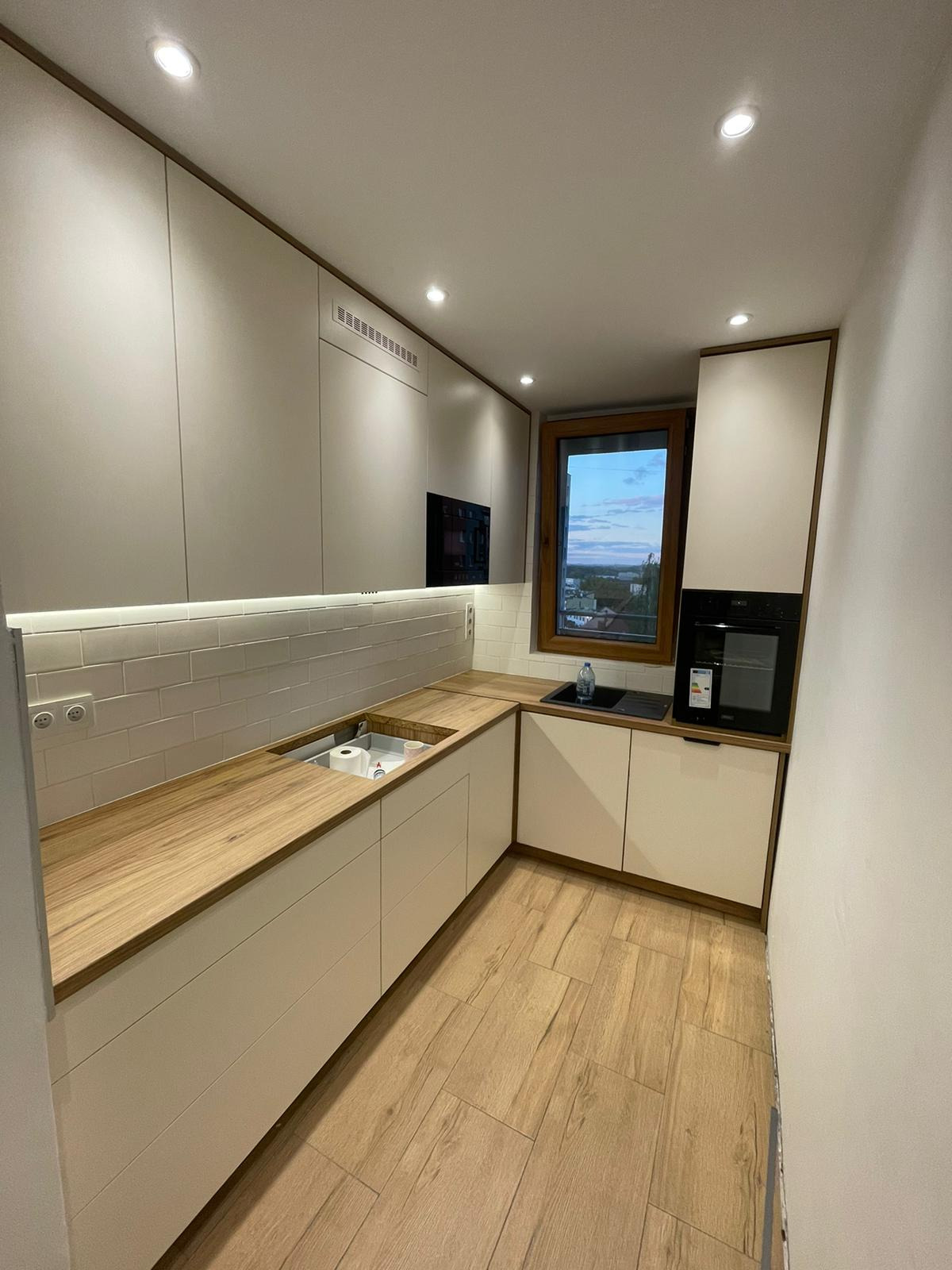 Jasna kuchnia z drewnianym blatem i oknem z widokiem na miasto. Minimalistyczny design, subtelne oświetlenie LED i płytki imitujące cegłę. Nowoczesne AGD w zabudowie.