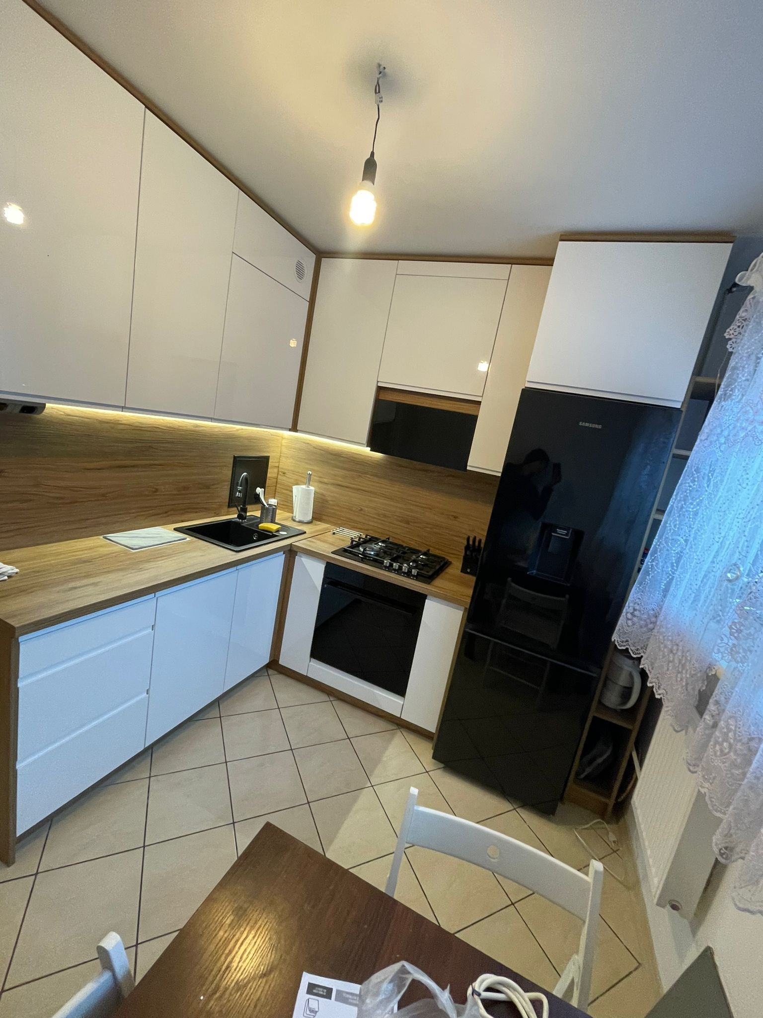 Nowoczesna kuchnia z białymi szafkami, drewnianym blatem i czarną lodówką Samsung. Oświetlenie LED pod szafkami górnymi. Widok na stół i krzesła.