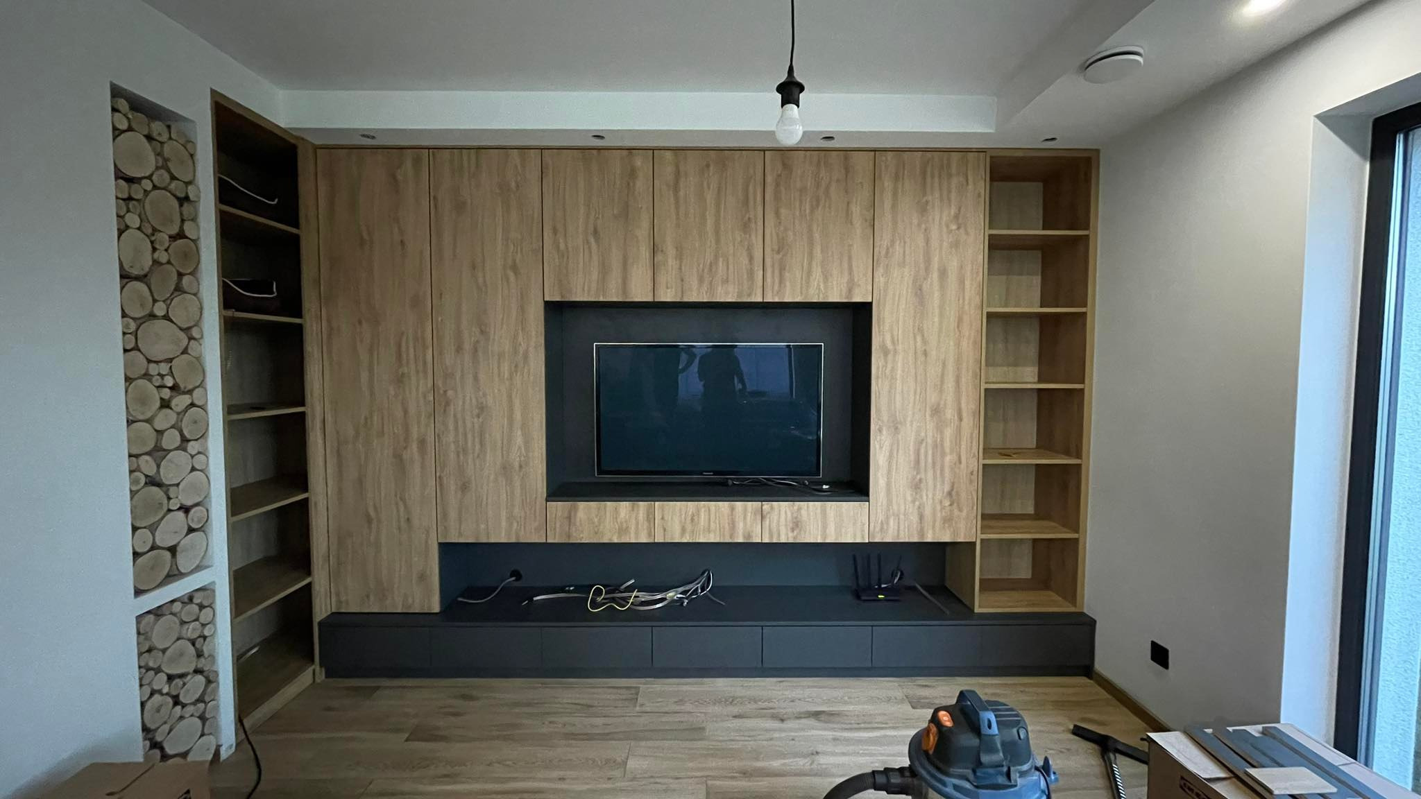 Drewniana zabudowa ściany z szafkami, półkami i miejscem na TV. Dekoracyjna wnęka z drewnianymi krążkami. Widoczny odkurzacz i narzędzia.