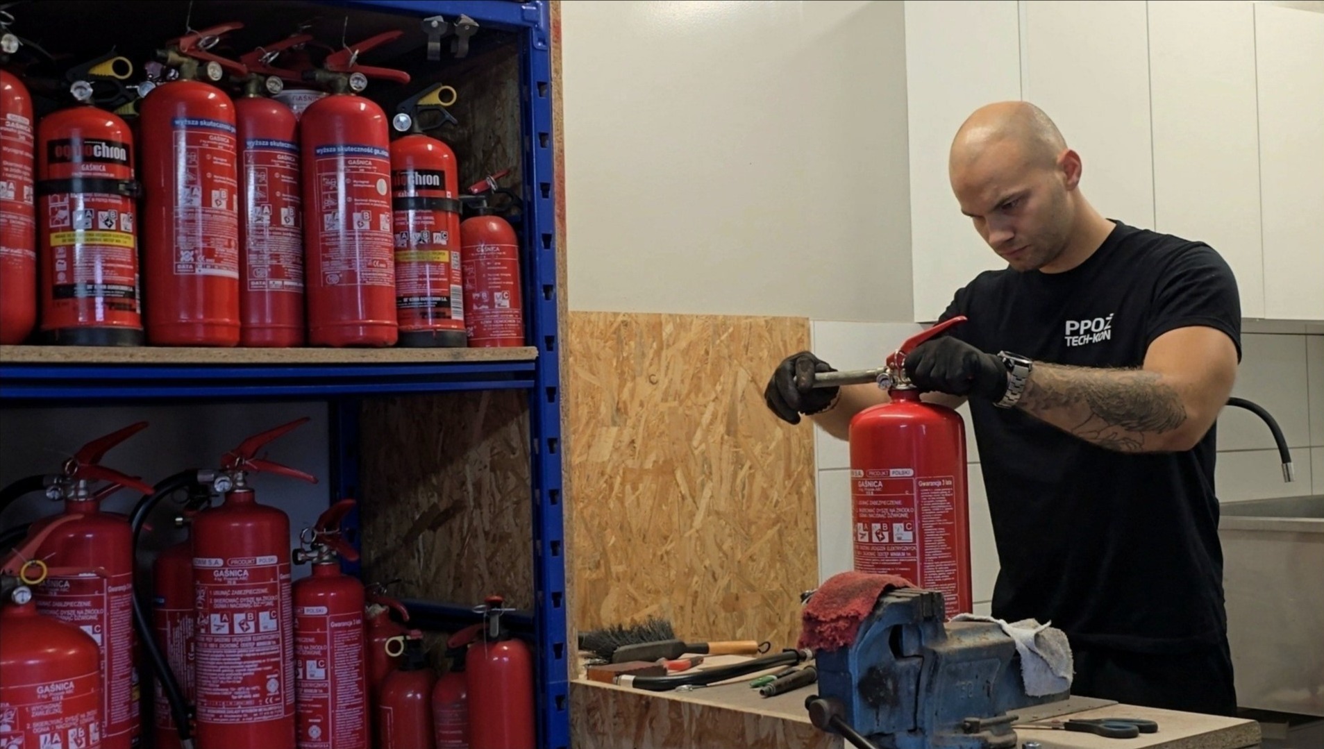 Przegląd gaśnic: mężczyzna w czarnych rękawicach serwisuje czerwoną gaśnicę, obok regał z zapasem, narzędzia na stole. Logo PPOŻ Tech-Kon na koszulce.