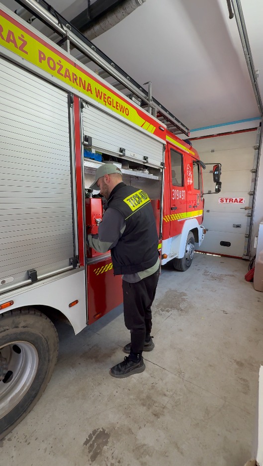 Strażak w kamizelce TECH-KAD otwiera schowek wozu strażackiego z napisem 'Straż Pożarna Węglewo' na tle otwartej bramy garażowej.