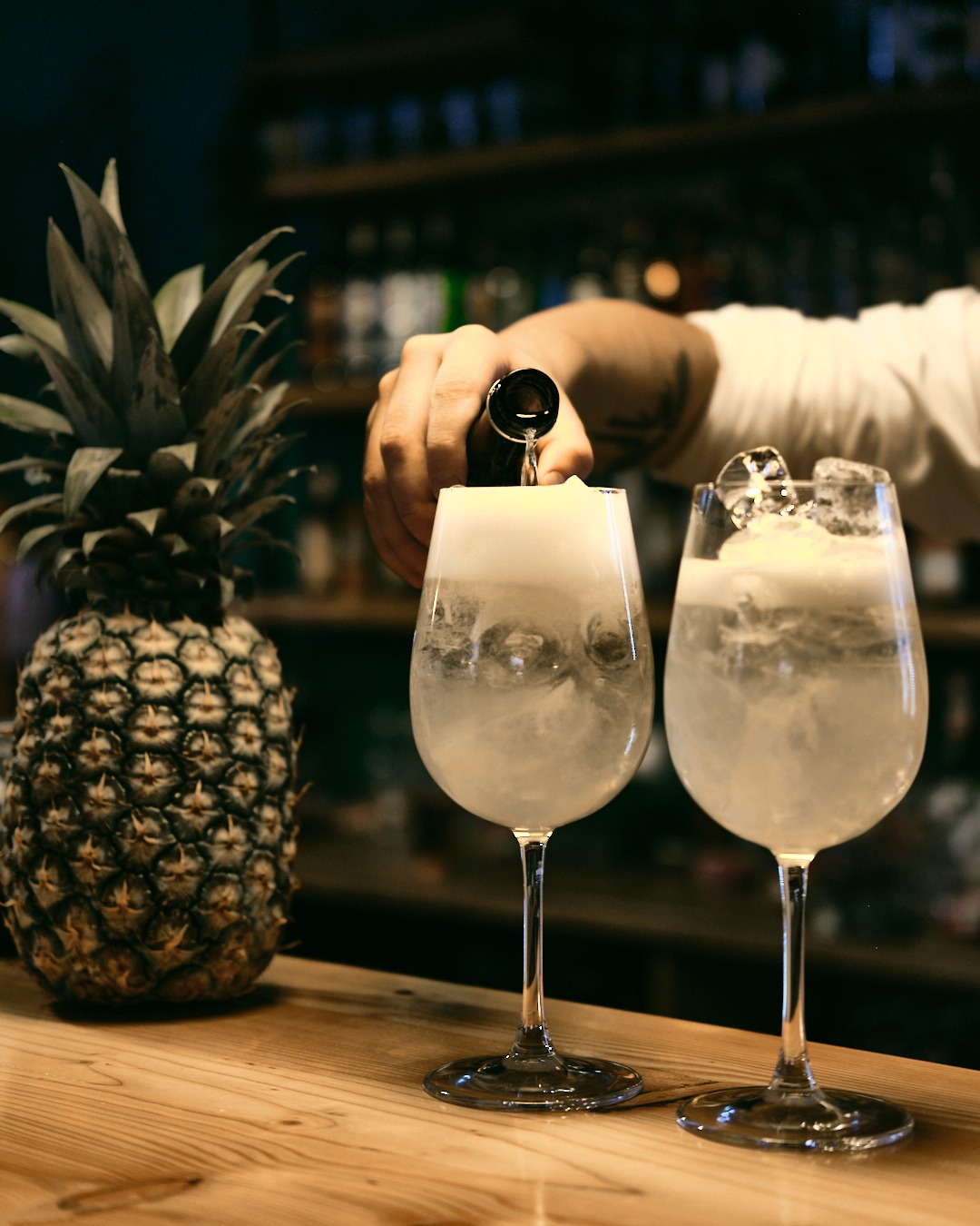 Barman nalewa drink do kieliszka z lodem, obok ananas na drewnianym blacie. W tle półki z butelkami. Ujęcie z bliska, ciepłe światło.