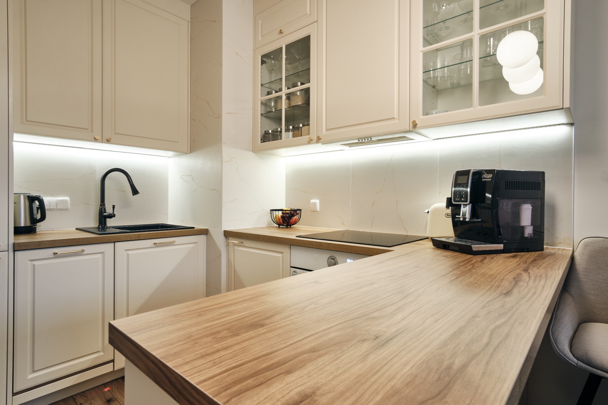 Jasna kuchnia z drewnianym blatem, czarną armaturą i sprzętami. Uwagę przykuwają przeszklone szafki górne z oświetleniem LED i dekoracyjnymi elementami.