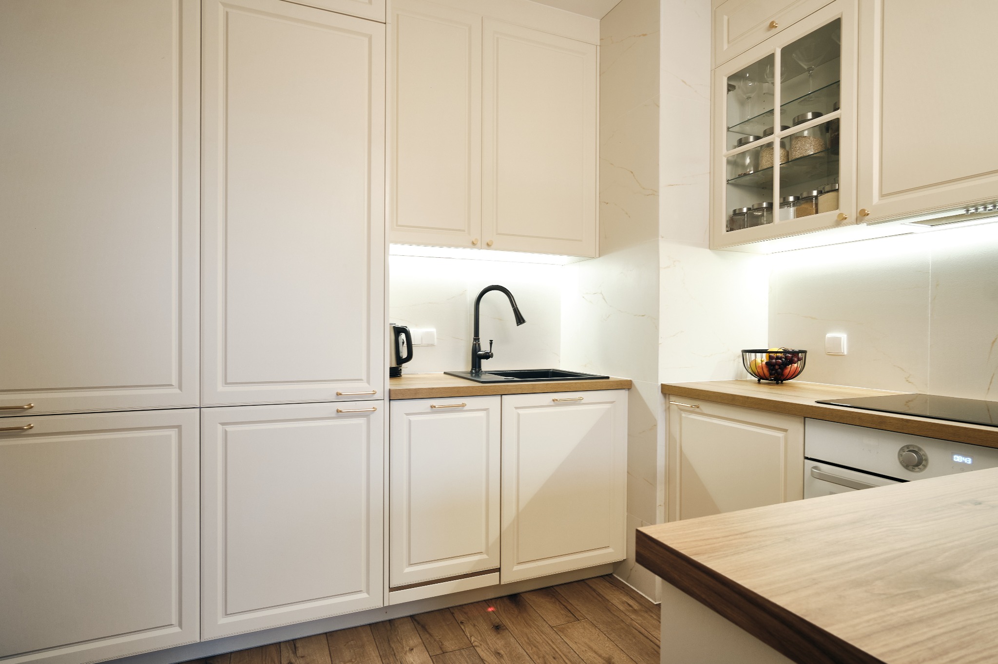Jasna kuchnia w klasycznym stylu: białe szafki z frezowanymi frontami, drewniany blat, czarna armatura i przeszklona witryna z dekoracyjnymi słoikami.