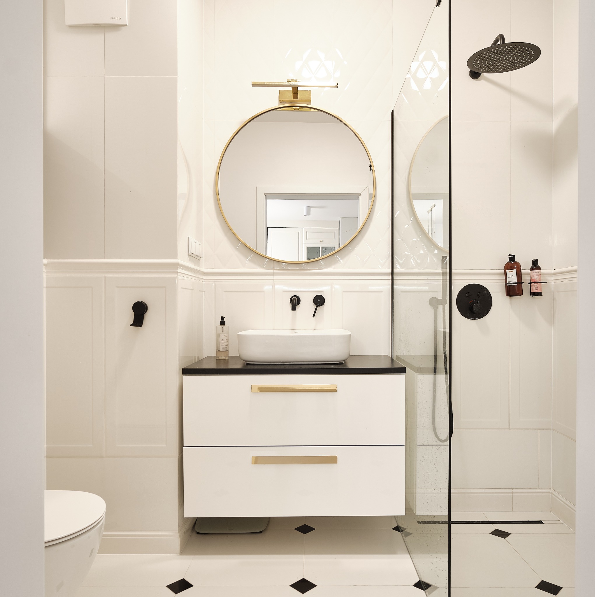 Elegancka, biała łazienka z czarnymi akcentami: okrągłe lustro w złotej ramie nad umywalką, kabina prysznicowa ze szklaną ścianką i czarną armaturą.