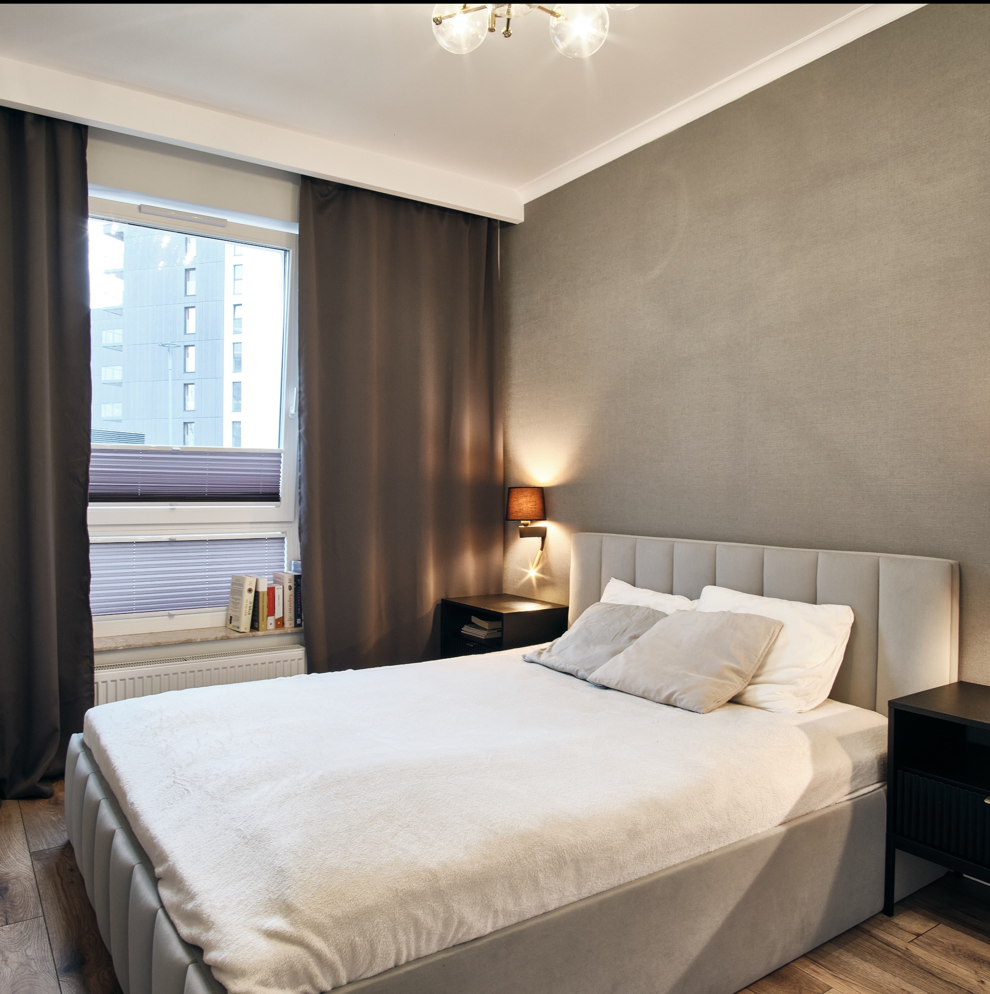 Neutralna sypialnia z szarymi ścianami, łóżkiem z tapicerowanym zagłówkiem i drewnianą podłogą. Okno z widokiem na budynek, zasłonięte brązowymi zasłonami.