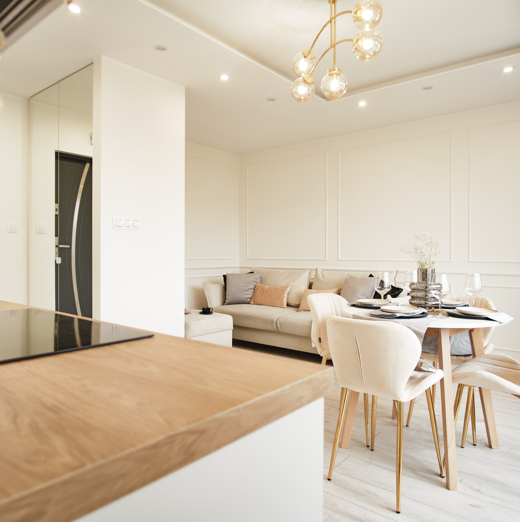 Jasne wnętrze: salon z sofą i jadalnią. Okrągły stół z nakryciem, krzesła z weluru, złoty żyrandol. Minimalistyczny design, neutralna kolorystyka, eleganckie wykończenie.
