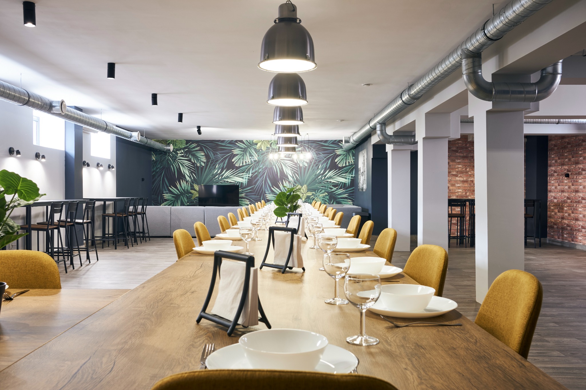 Długi stół z nakryciami i żółtymi krzesłami w przestronnym wnętrzu z muralem z motywem roślinnym i widoczną instalacją wentylacyjną pod sufitem.