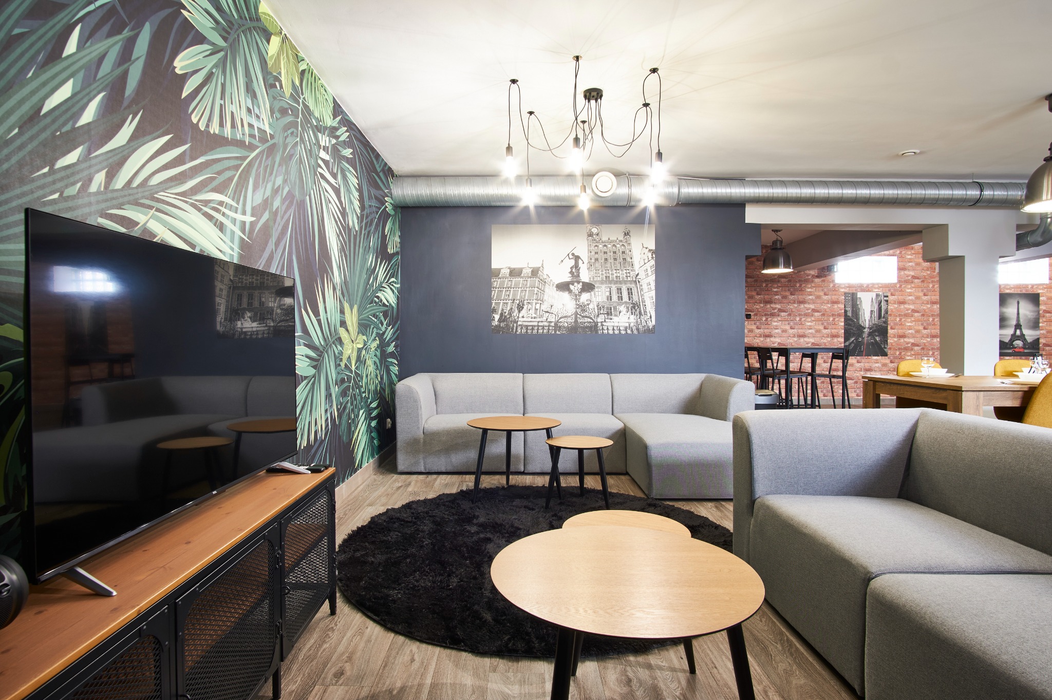 Nowoczesne wnętrze z szarą sofą modułową, okrągłym czarnym dywanem i stolikami kawowymi. Na ścianie fototapeta z motywem roślinnym i czarno-biała fotografia.