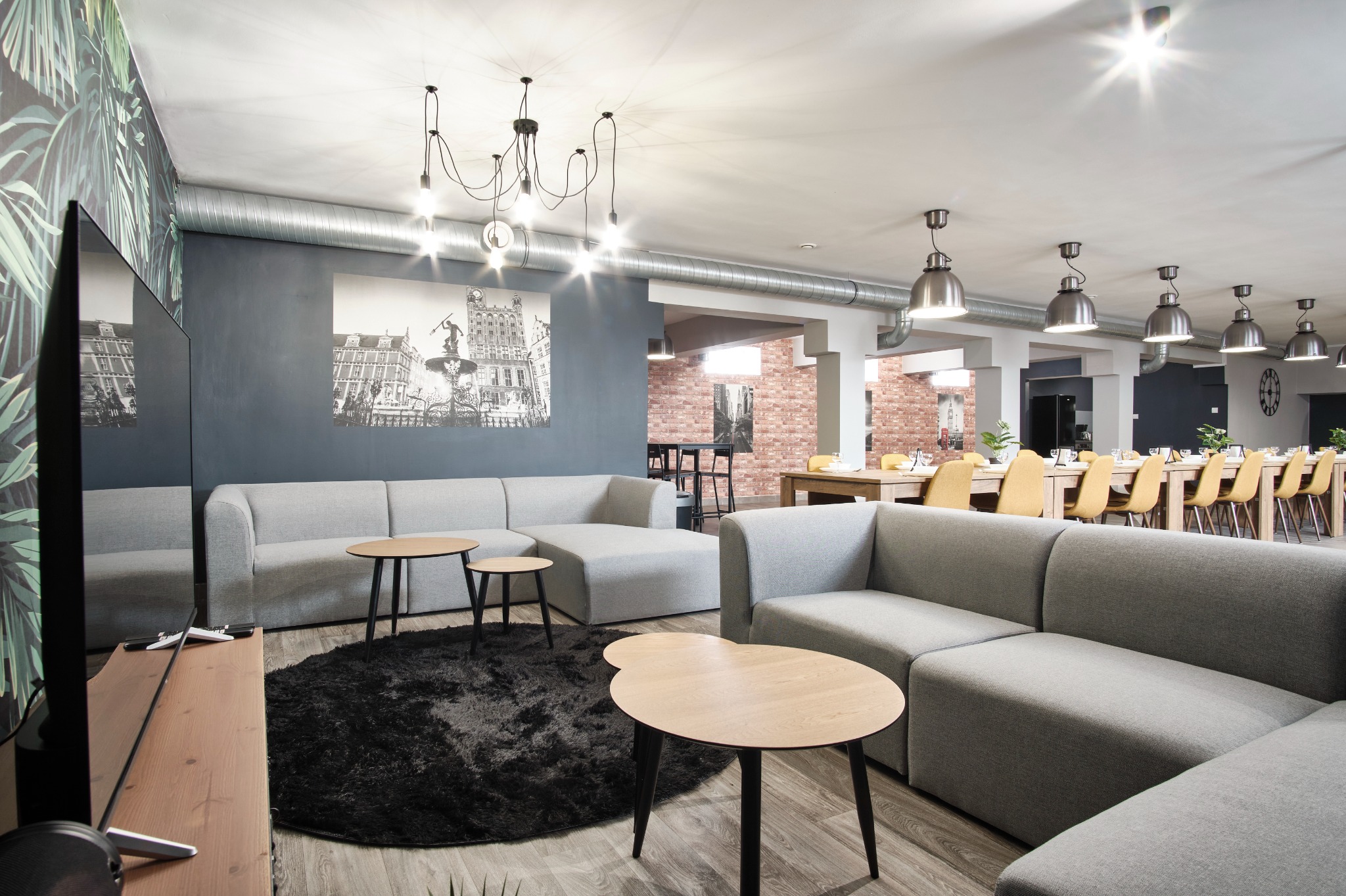 Przestronne wnętrze biurowe z szarymi sofami modułowymi, drewnianymi stolikami i czarnym dywanem. W tle długa jadalnia z żółtymi krzesłami i ceglana ściana.