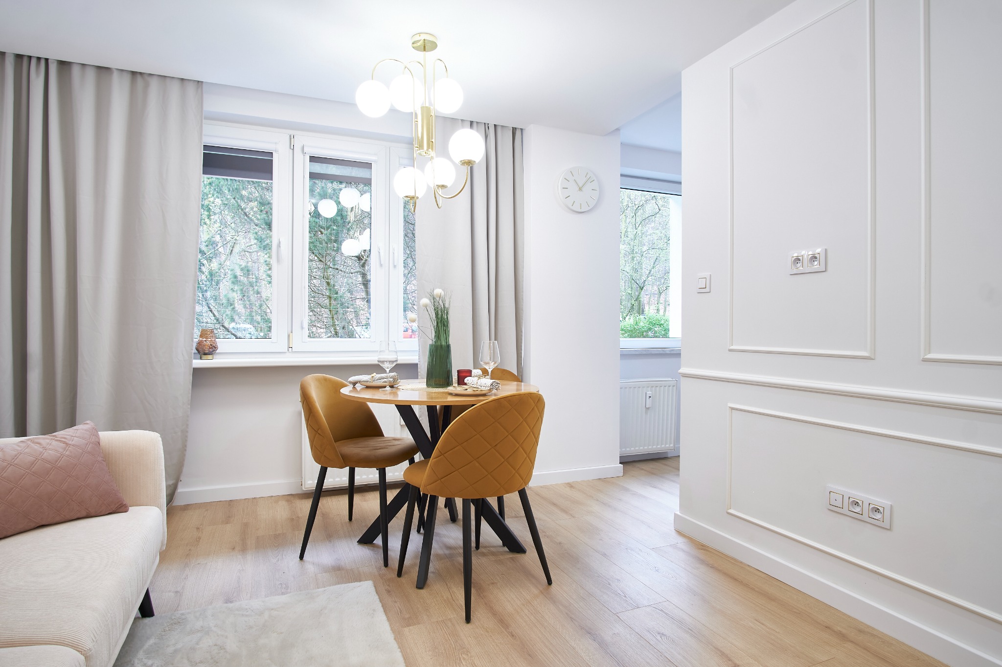 Jasne wnętrze jadalni z okrągłym stołem i musztardowymi krzesłami na tle okna z widokiem na zieleń. Minimalistyczny design, drewniana podłoga i dekoracyjne listwy ścienne.