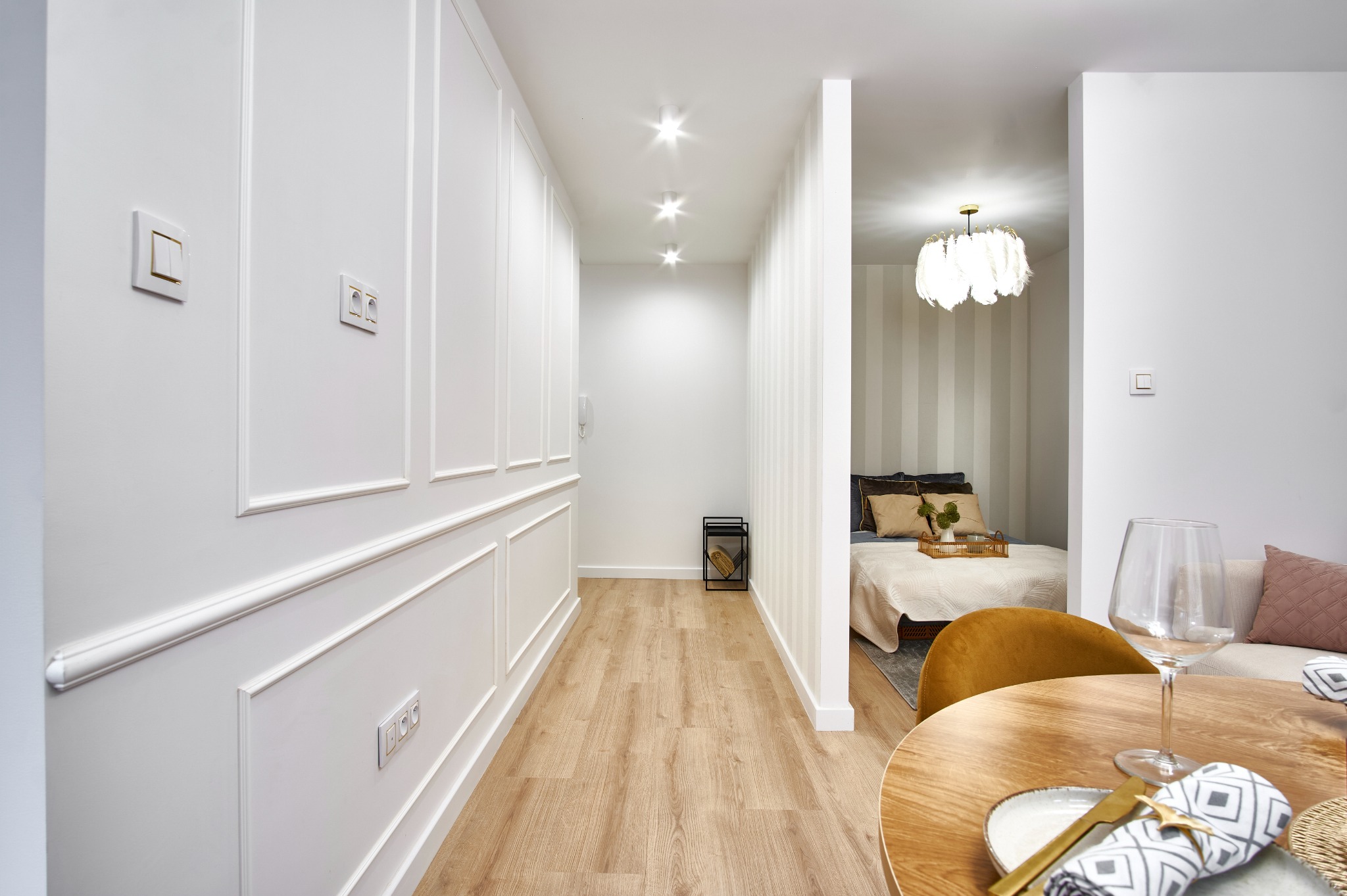 Jasne wnętrze mieszkania z drewnianą podłogą, białymi ścianami z dekoracyjnymi listwami i widokiem na sypialnię z okrągłym stołem jadalnym na pierwszym planie.
