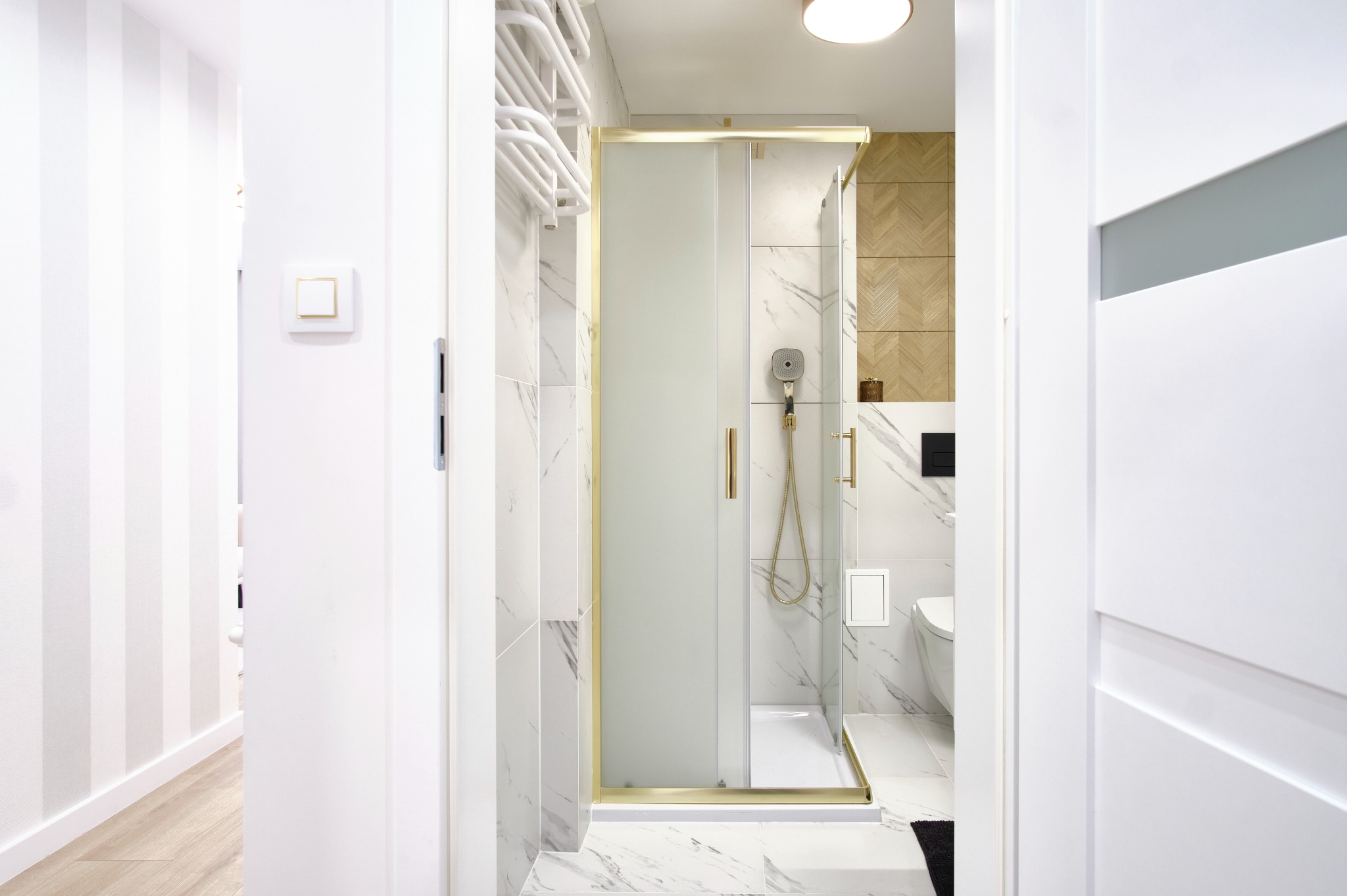 Nowoczesna łazienka z kabiną prysznicową ze złotymi detalami, marmurowymi płytkami i drewnianą mozaiką, widziana z perspektywy wejścia.